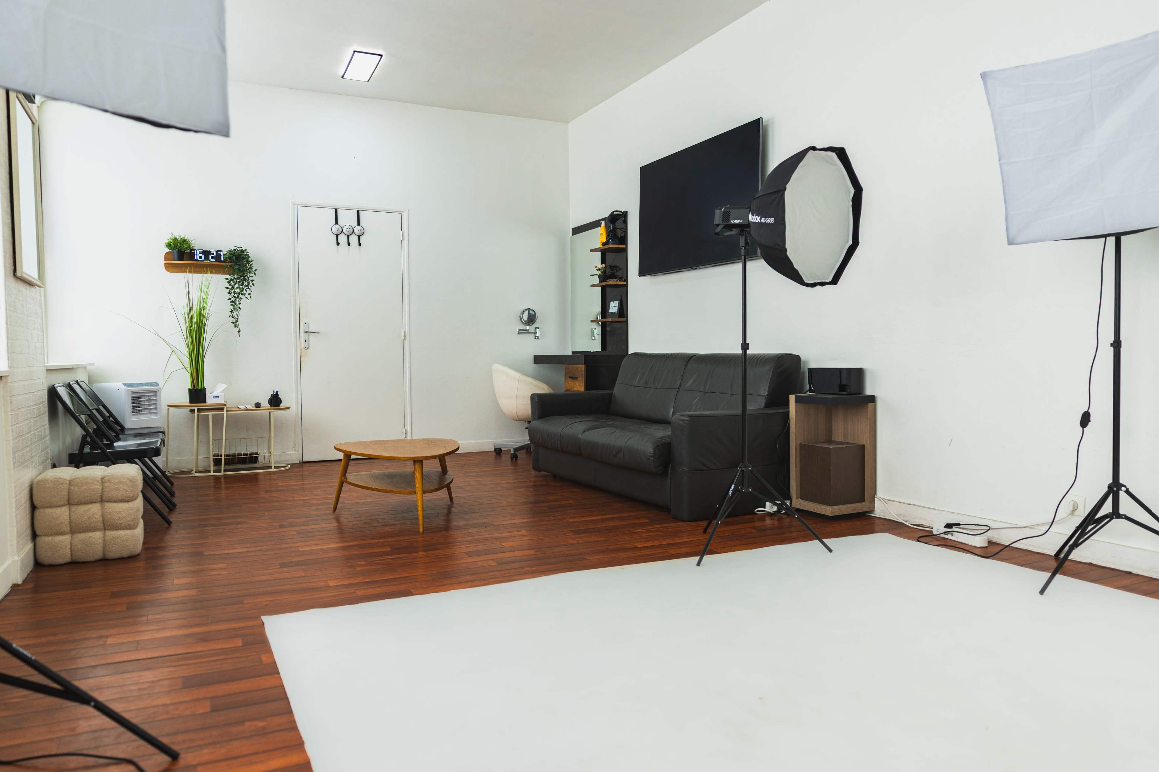 The image shows a well-lit studio space featuring a black sofa, a round wooden table, and photography equipment, including softboxes and a backdrop.
