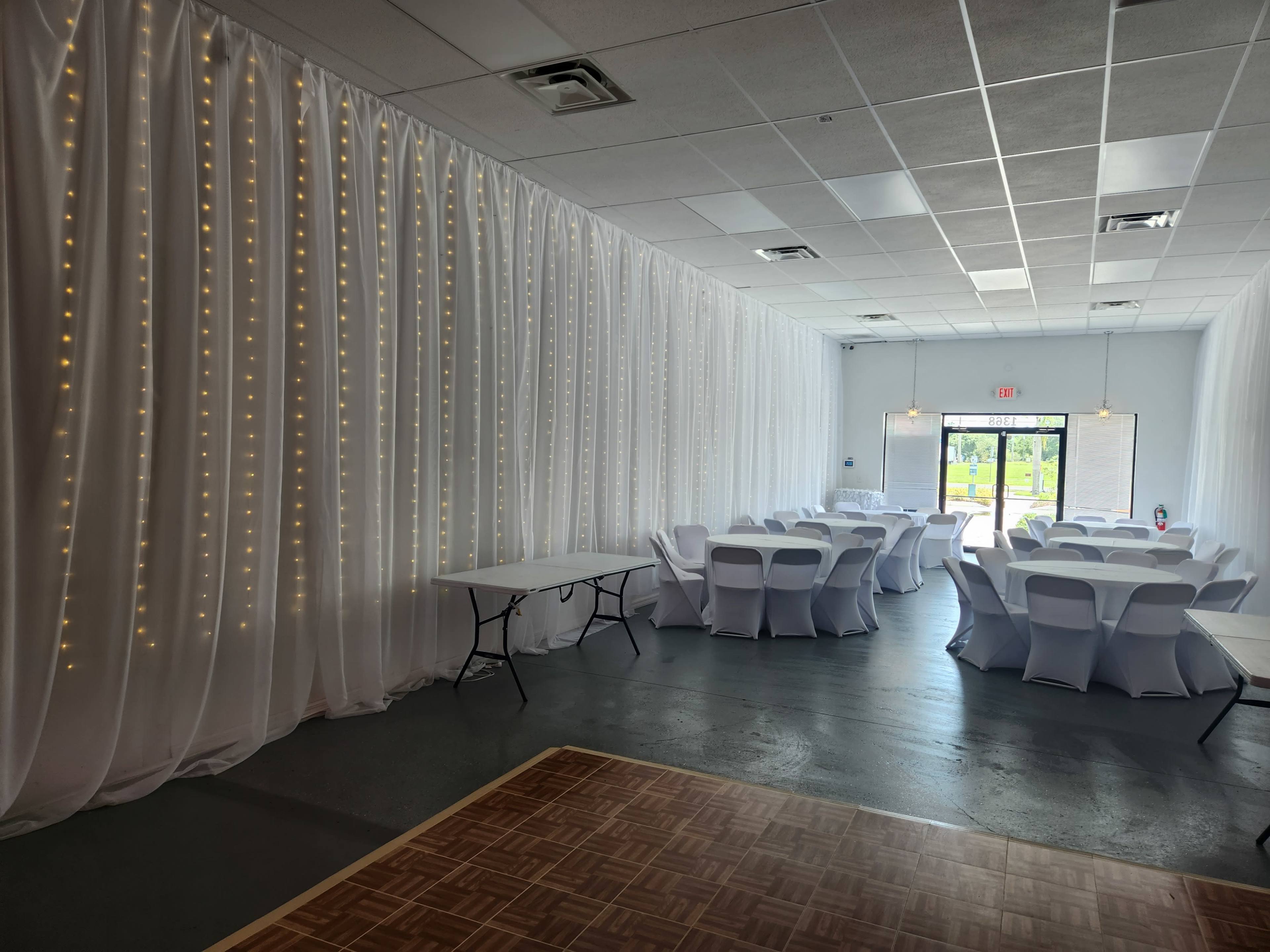 A decorated event space with white draped curtains, string lights, and tables arranged with white covers.