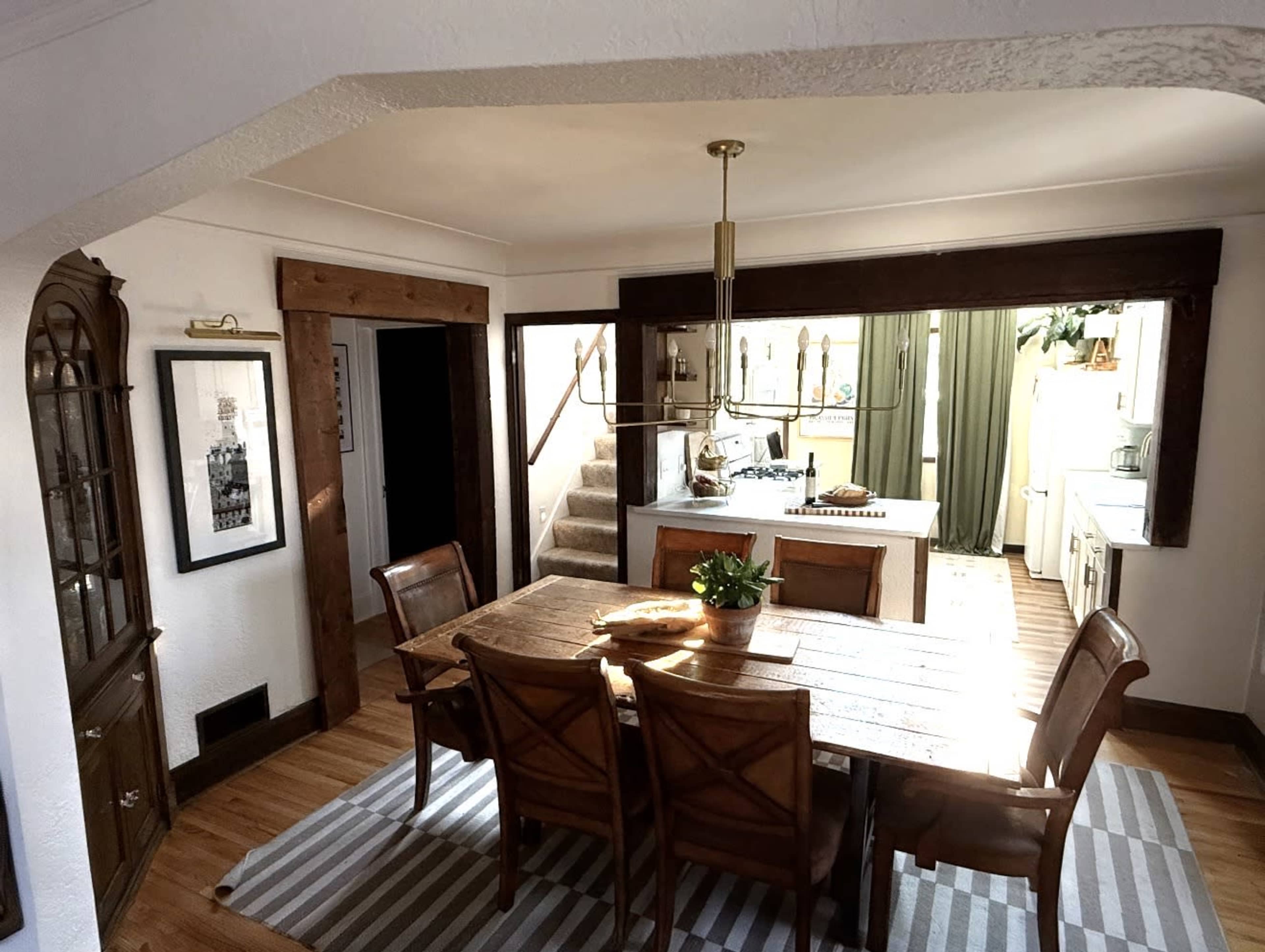 A wooden dining table is surrounded by chairs in a cozy dining room, with a kitchen visible in the background and green curtains hanging in the windows.