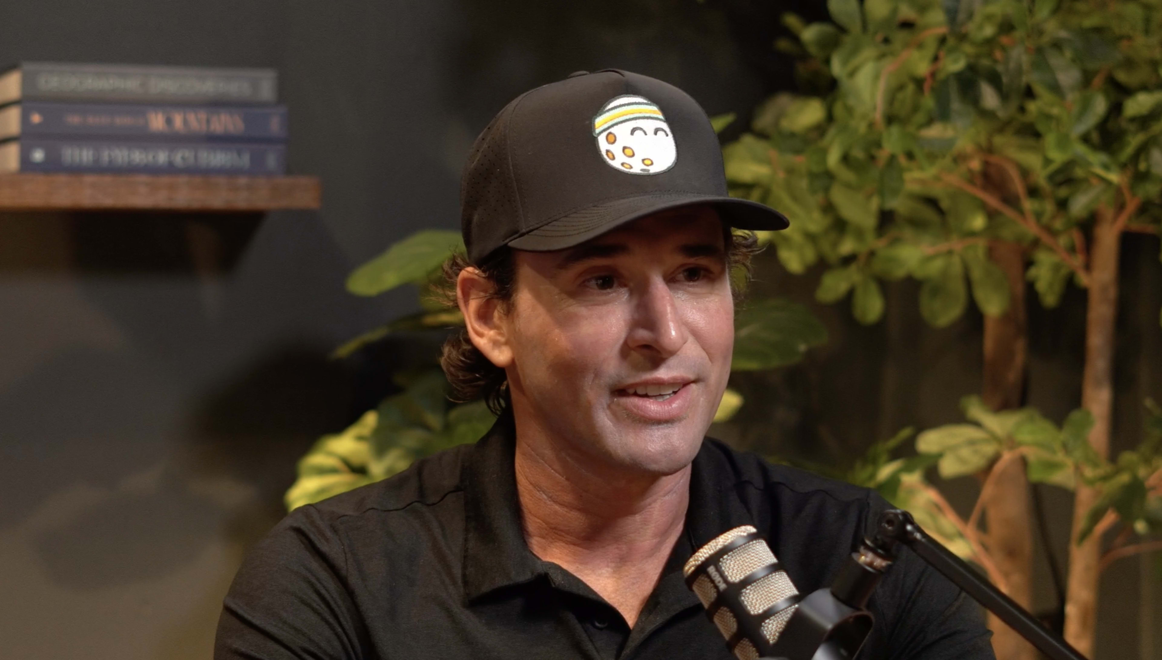 A man in a black cap with a smiling graphic is speaking in front of a microphone and surrounded by plants.