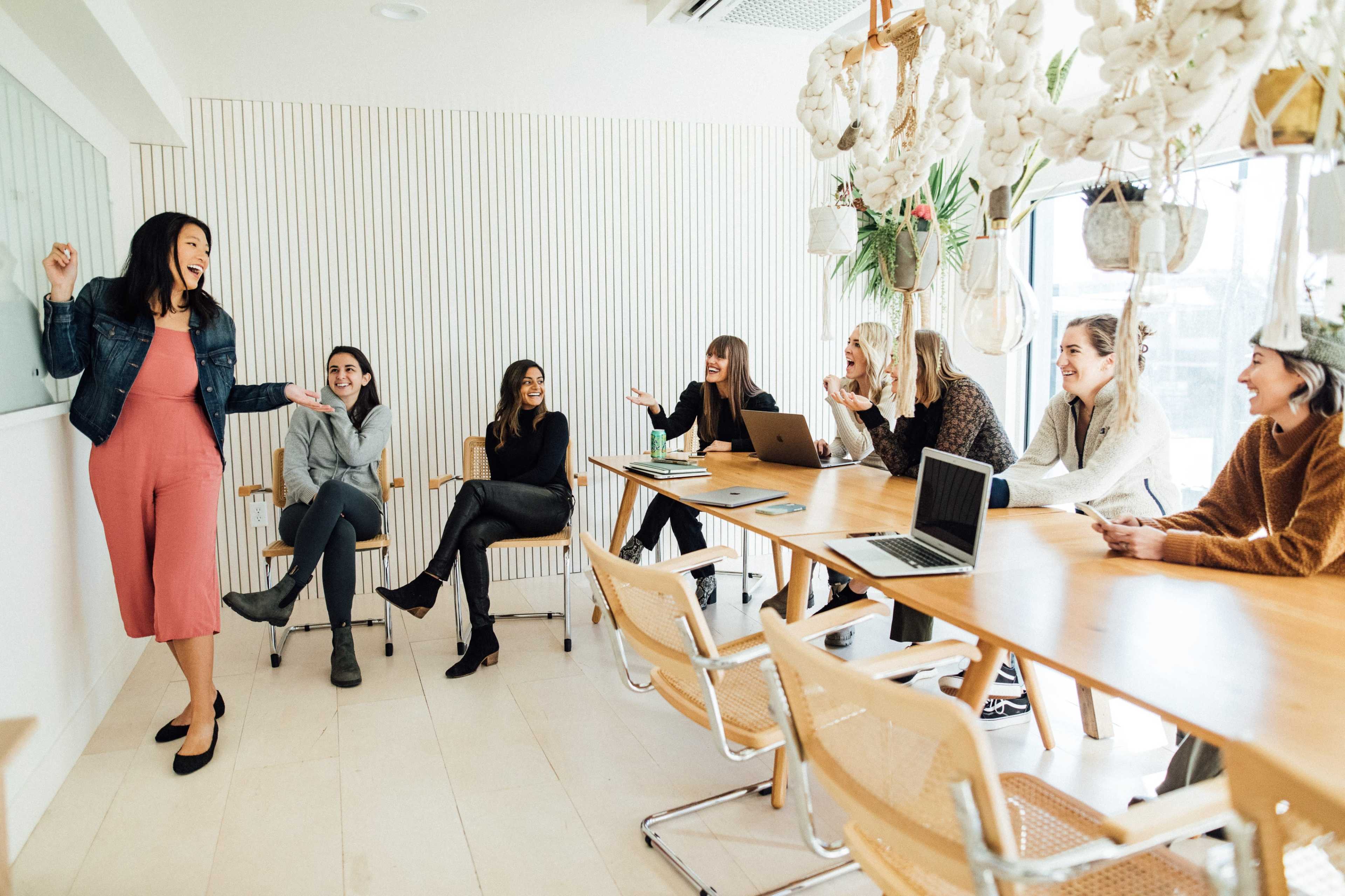 A diverse group of women sits around a large table in a bright meeting room, engaged in a discussion while one woman stands at a whiteboard, presenting.