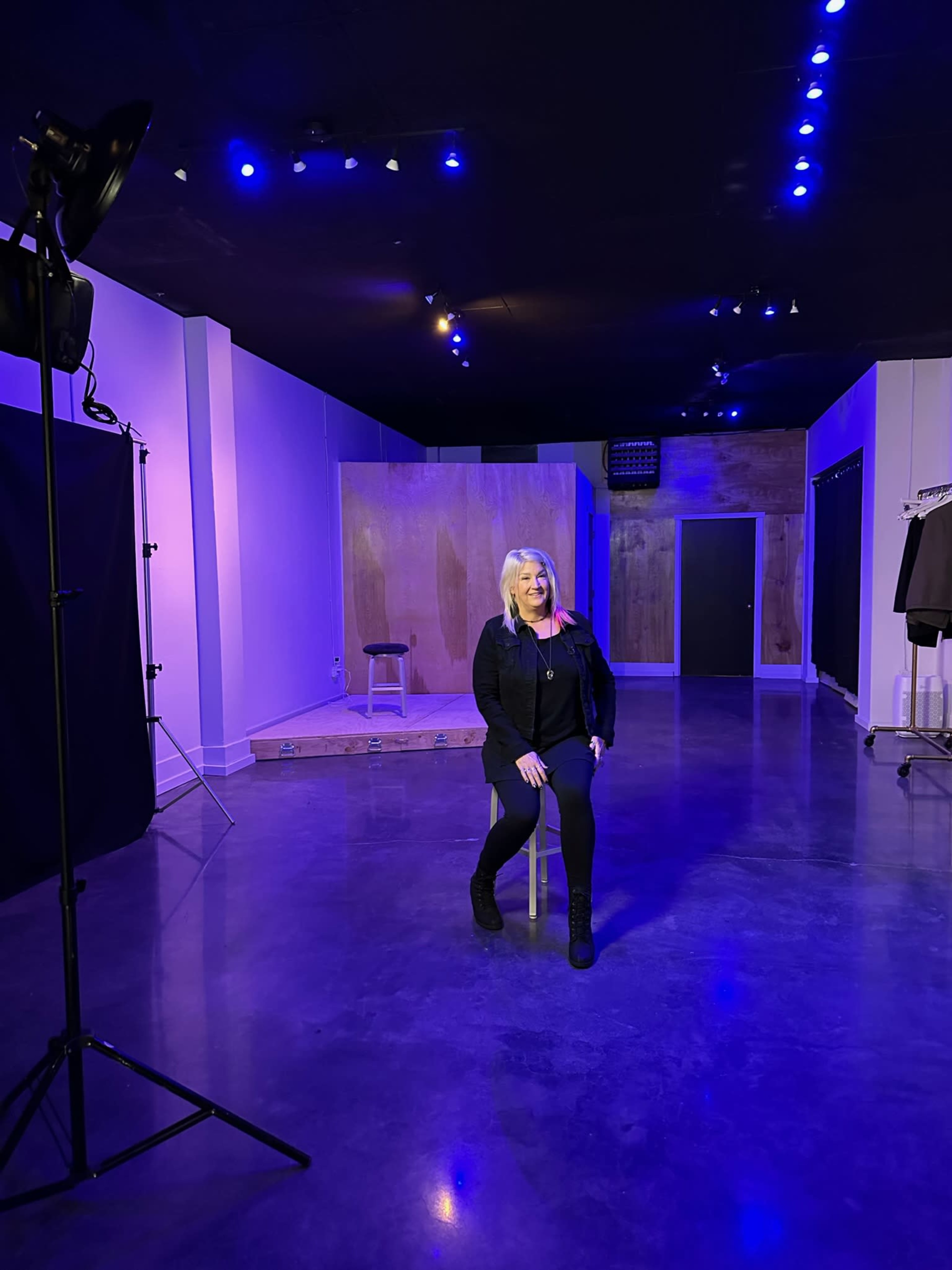 A woman sits on a stool in an empty studio with purple lighting.
