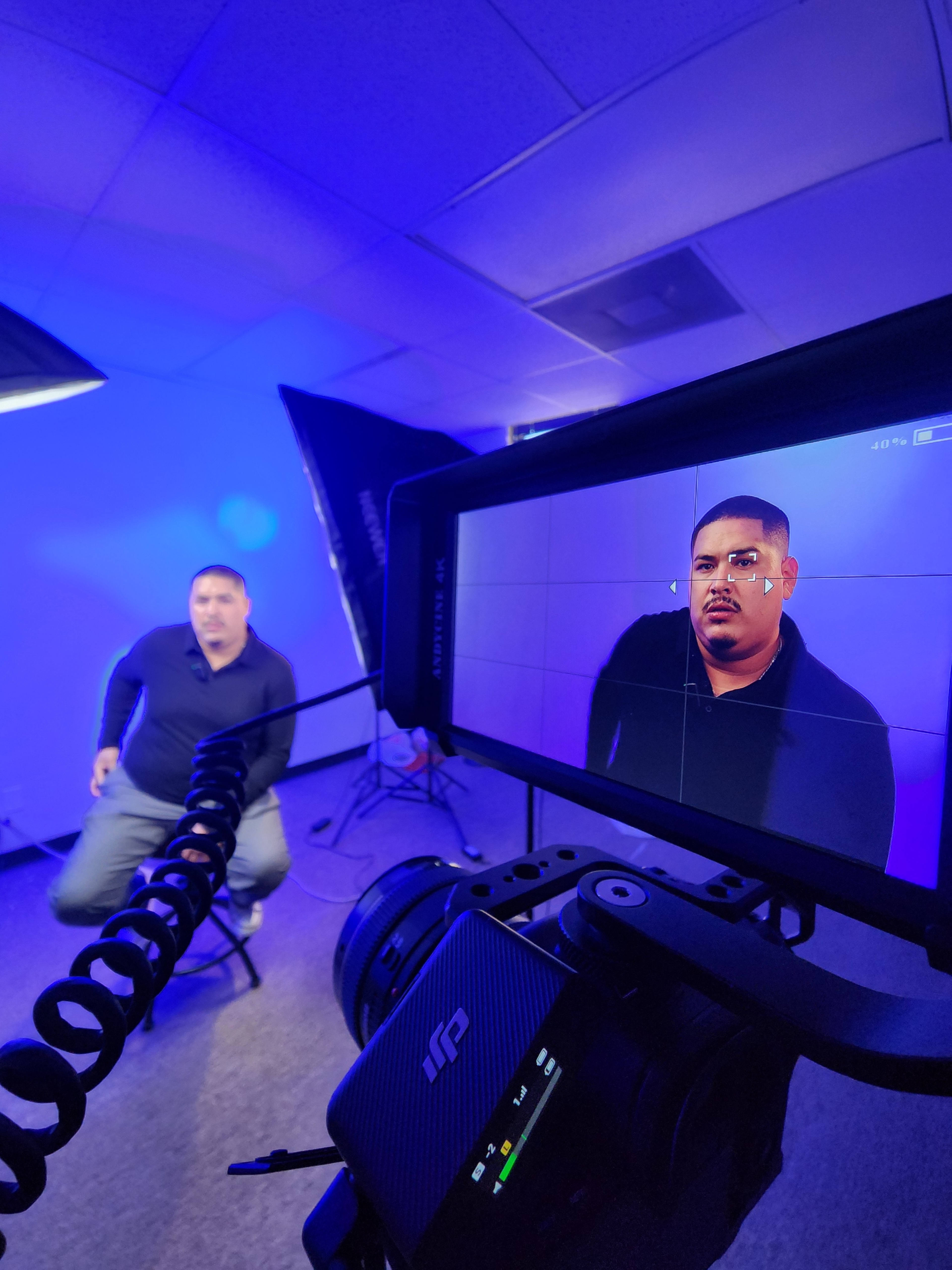 A man sits on a stool in a lit studio while being recorded by a camera positioned in front of him.