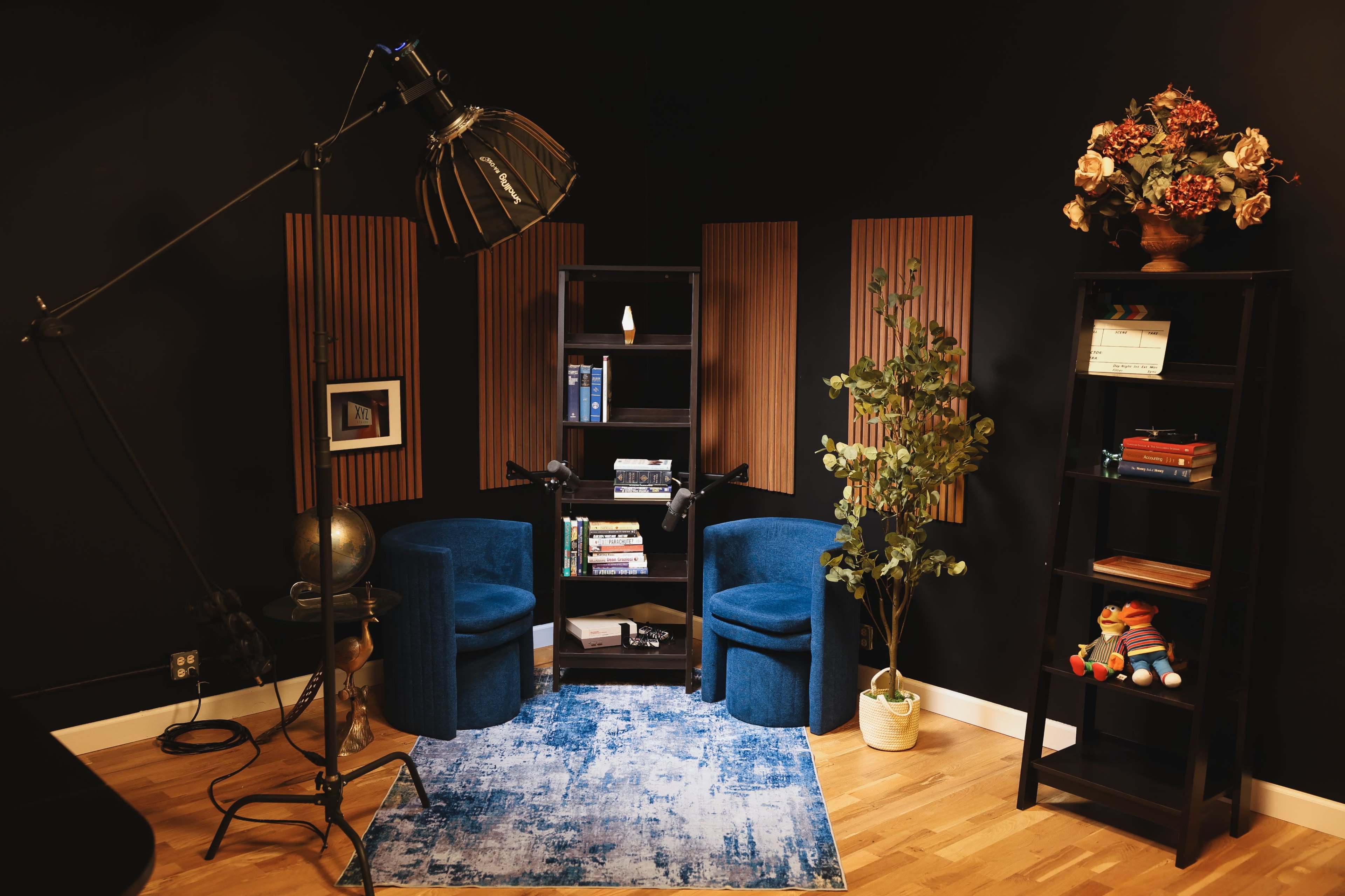 The image shows a cozy corner with two blue armchairs, a bookshelf filled with books, decorative plants, and a large lamp against a dark wall.