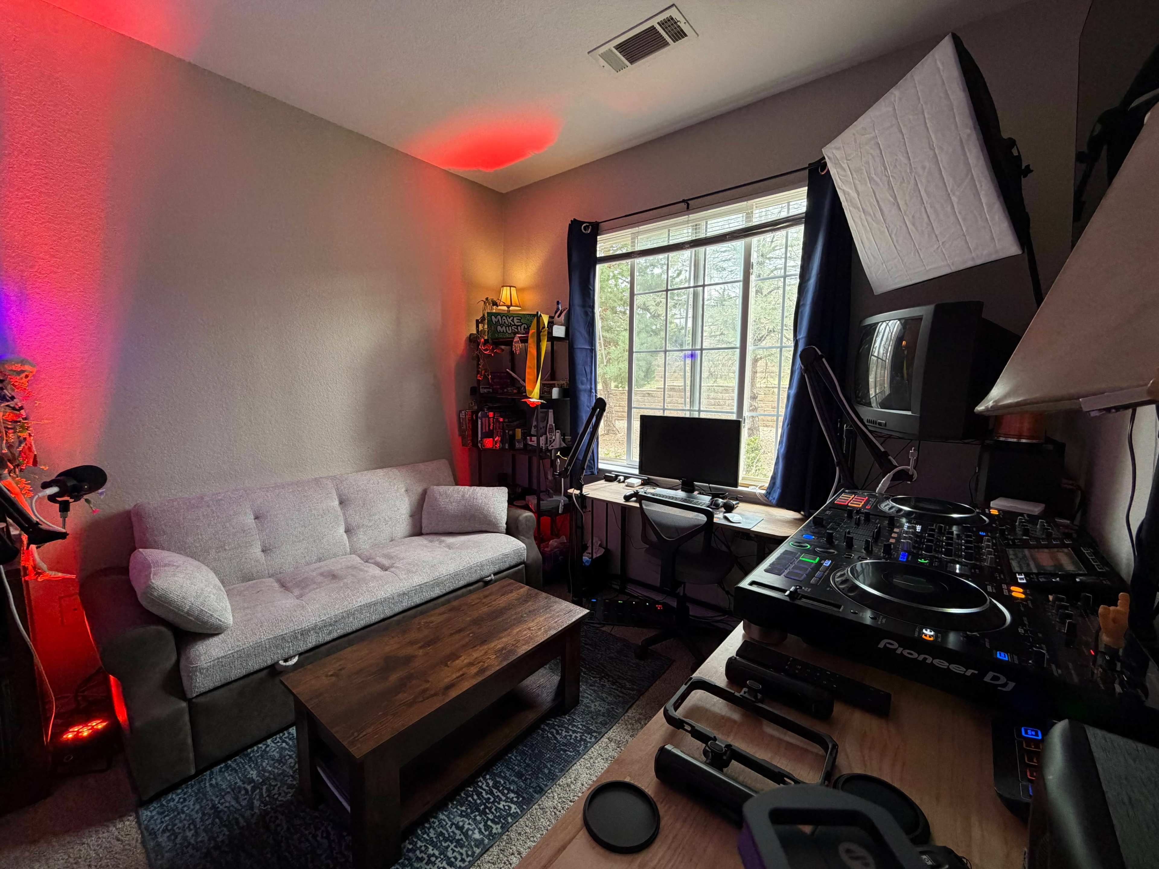 The image depicts a small room with a gray couch, a wooden coffee table, a computer setup, and DJ equipment, illuminated by soft red and natural light from a window.