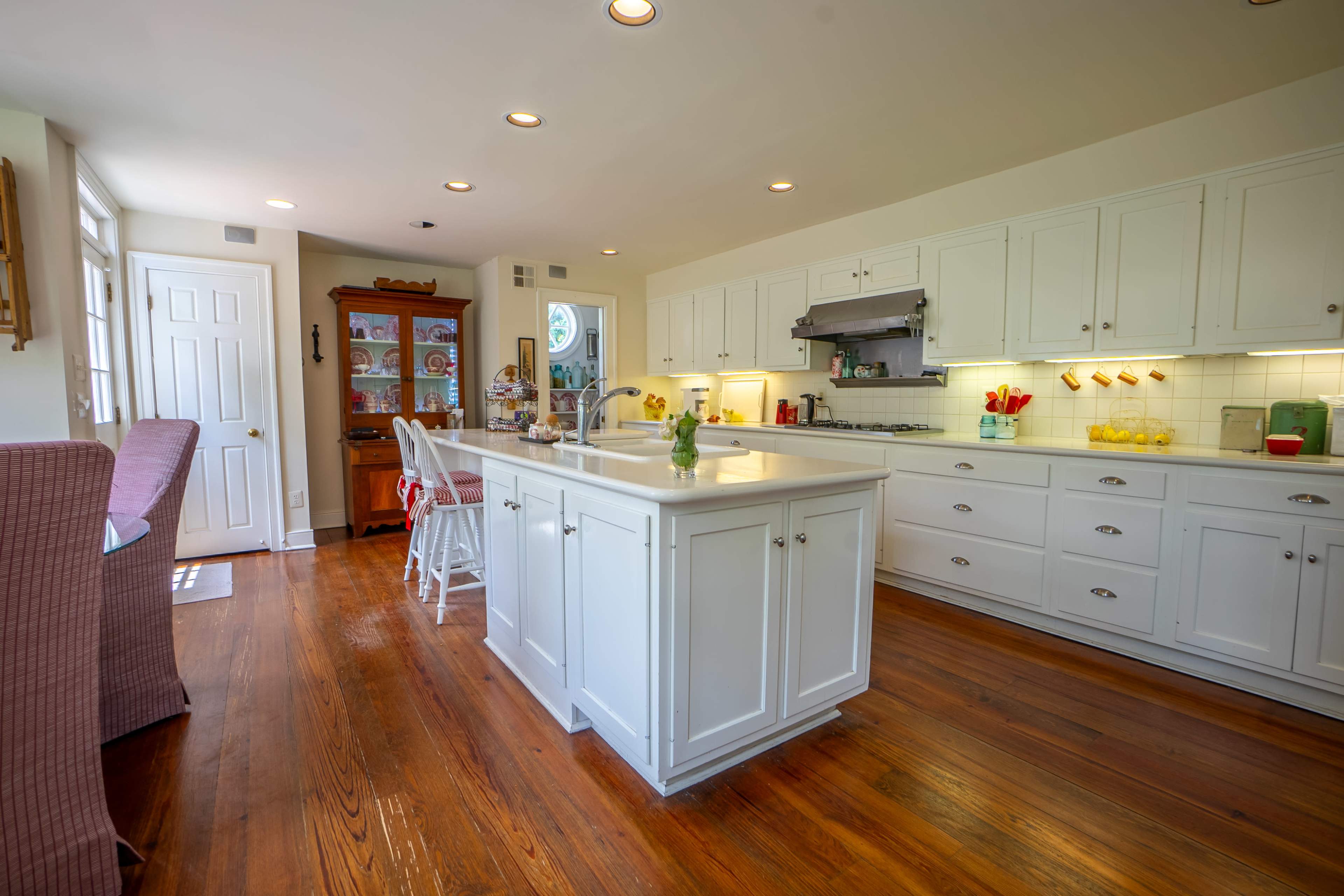 The image shows a bright kitchen with white cabinets, a large island, and wooden flooring, featuring a seating area and a variety of kitchen appliances.