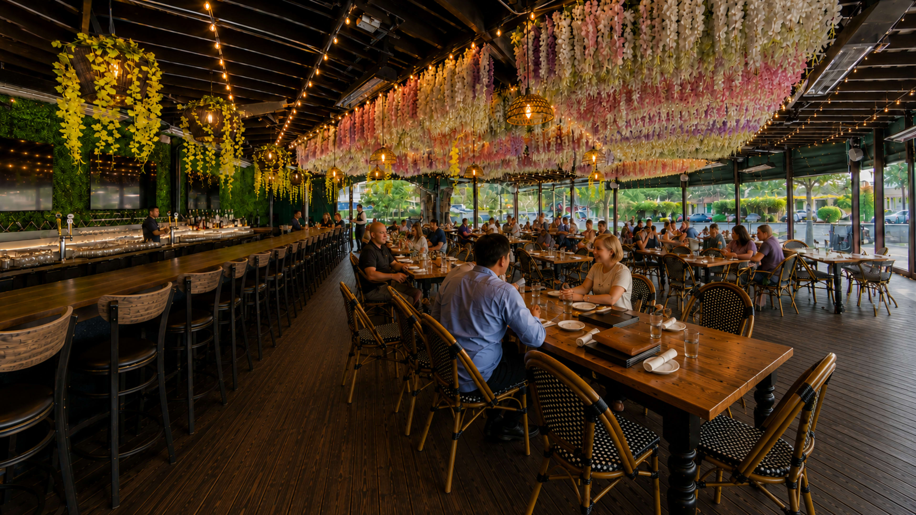 The image shows a vibrant restaurant interior with a long bar, numerous tables, and colorful floral decorations hanging from the ceiling.