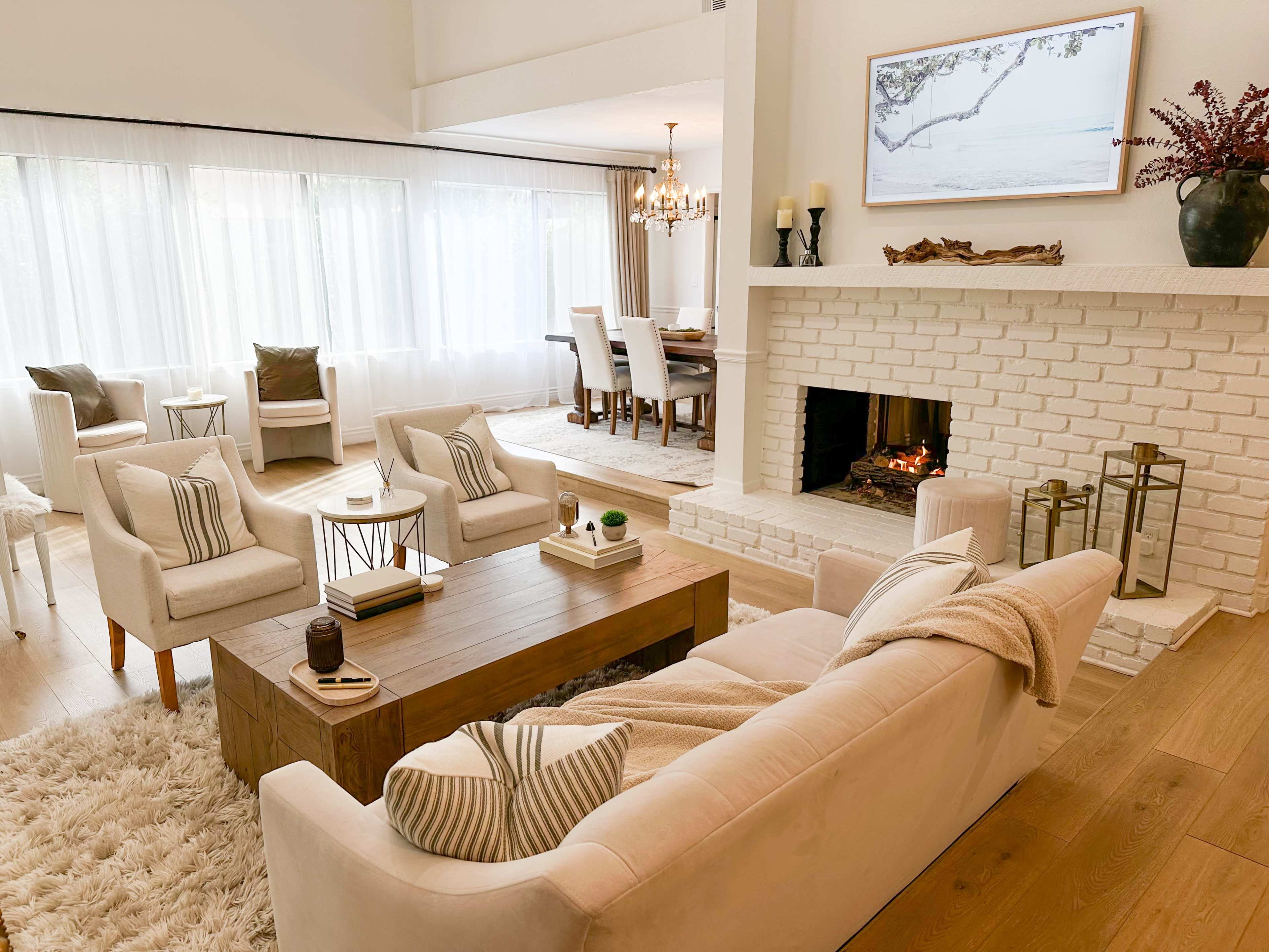 The image shows a stylish living room featuring a light-colored sofa, two armchairs, a wooden coffee table, and a fireplace, with a dining area in the background.