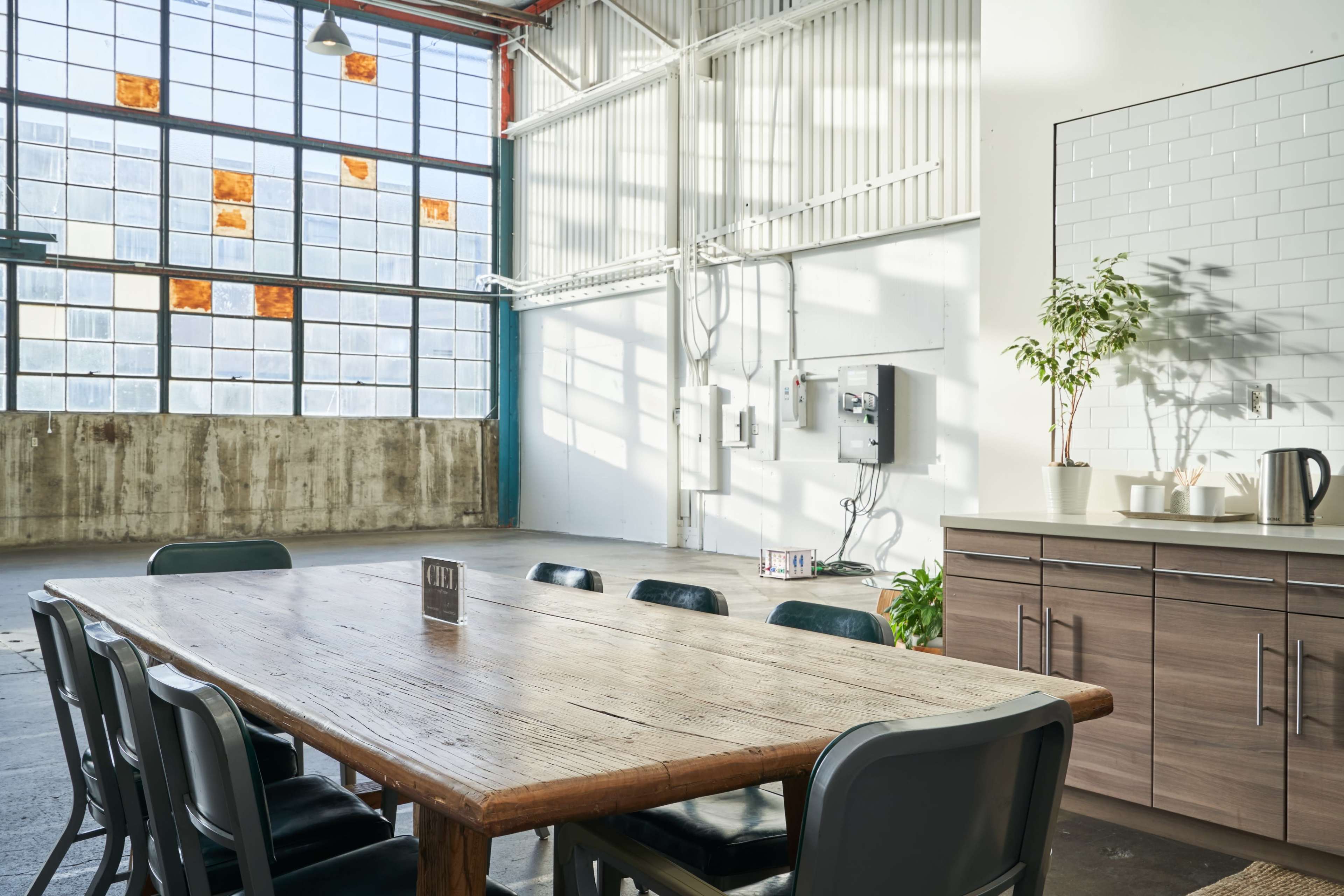 A large wooden table with black chairs is positioned in a spacious, brightly lit room featuring large industrial windows and a minimalist kitchen area.