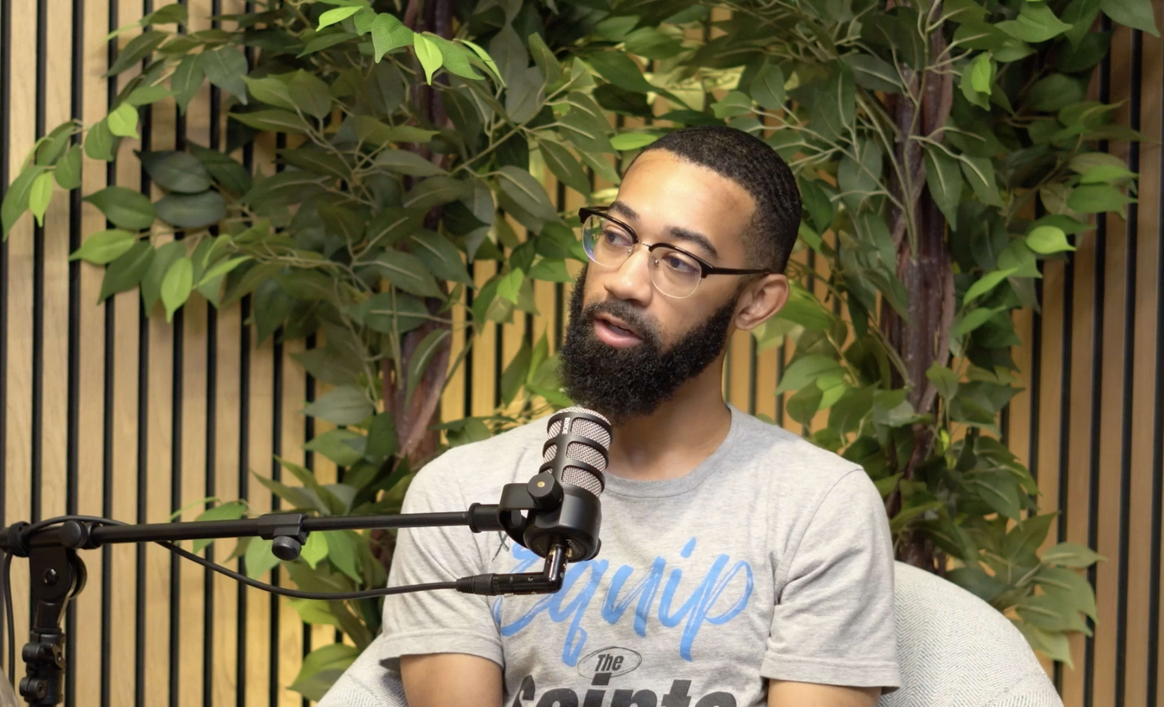 A man with a beard and glasses sits in front of a microphone amid green foliage.