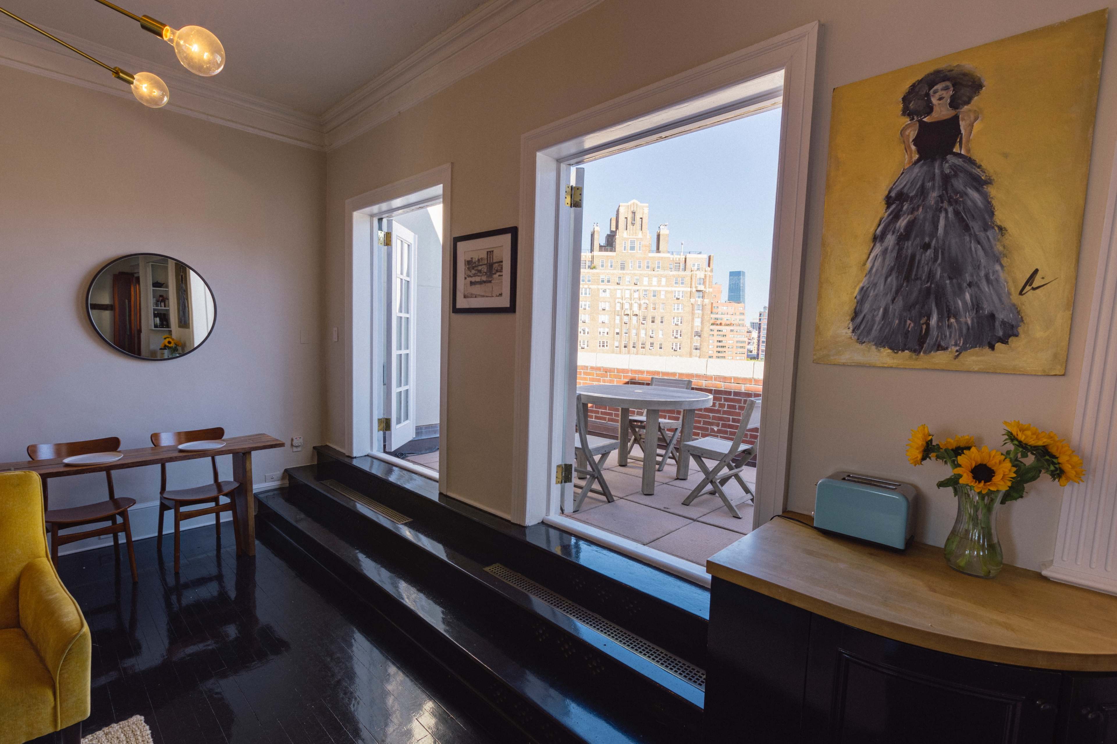The image shows a bright room with two open doors leading to a balcony, featuring a round mirror, a painting of a woman in a stylish dress, a yellow armchair, and a small table with chairs.