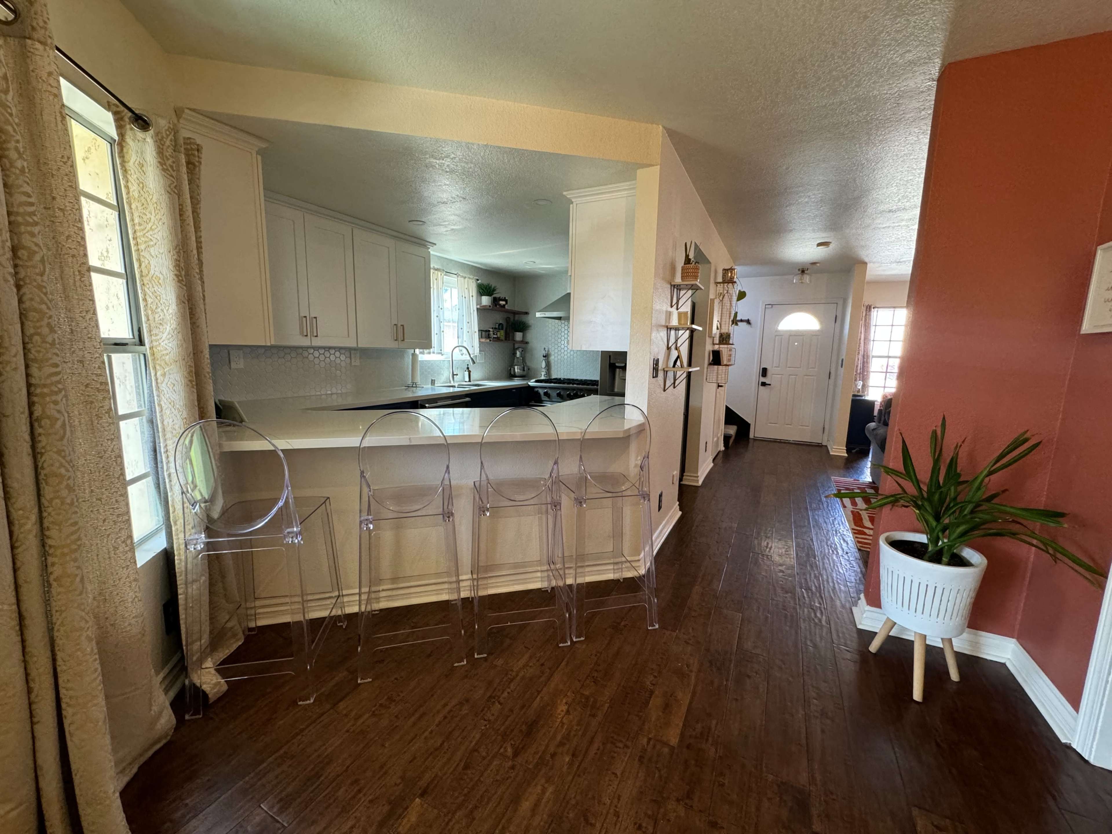 The image shows a modern kitchen area with transparent bar stools, adjoining a hallway leading to a front door, and a potted plant in the corner.