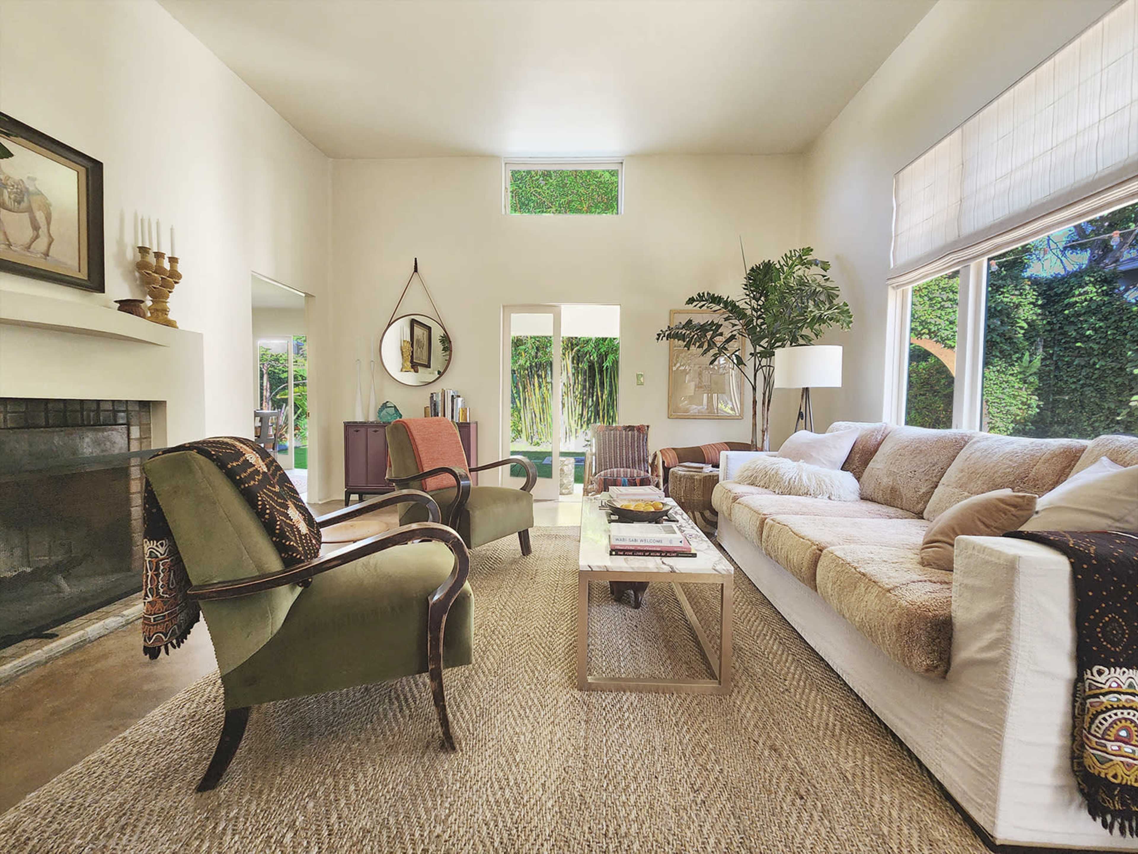 A bright living room with a white sofa, two armchairs, and large windows overlooking a green outdoor space.