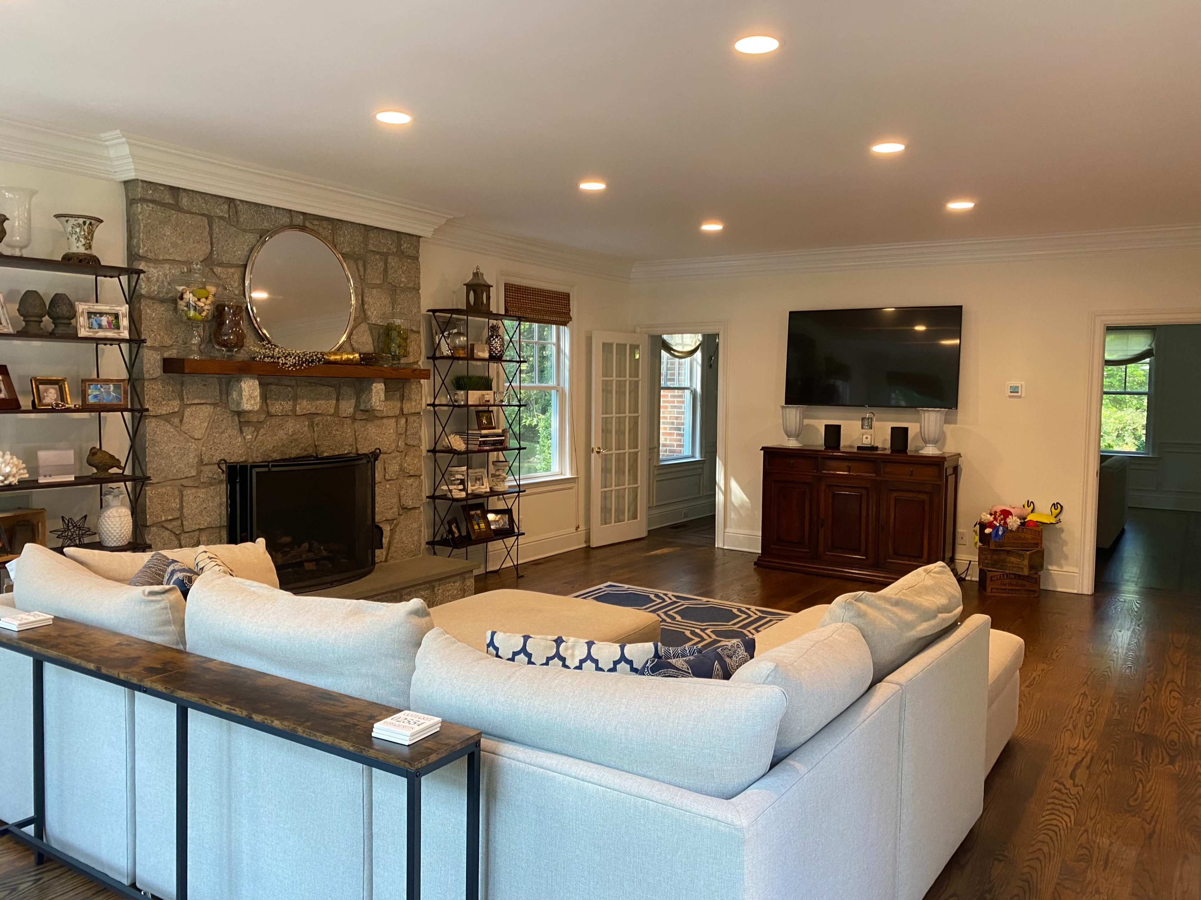 The image shows a bright living room with a large sectional sofa, a stone fireplace with a circular mirror above it, built-in shelves, and a wall-mounted television.