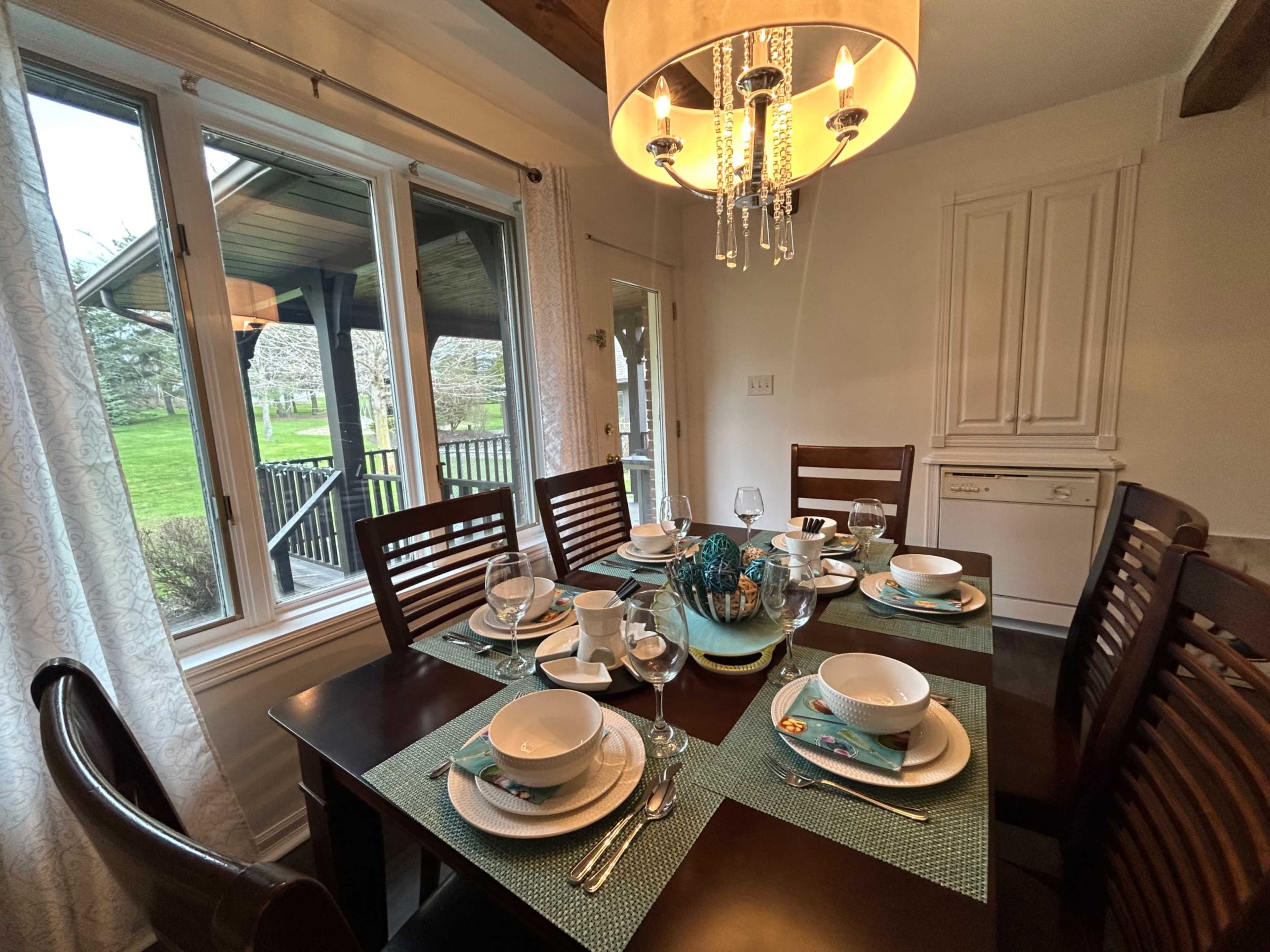 A dining table is set with white plates and silverware, surrounded by wooden chairs, and overlooks a green outdoor area through large windows.
