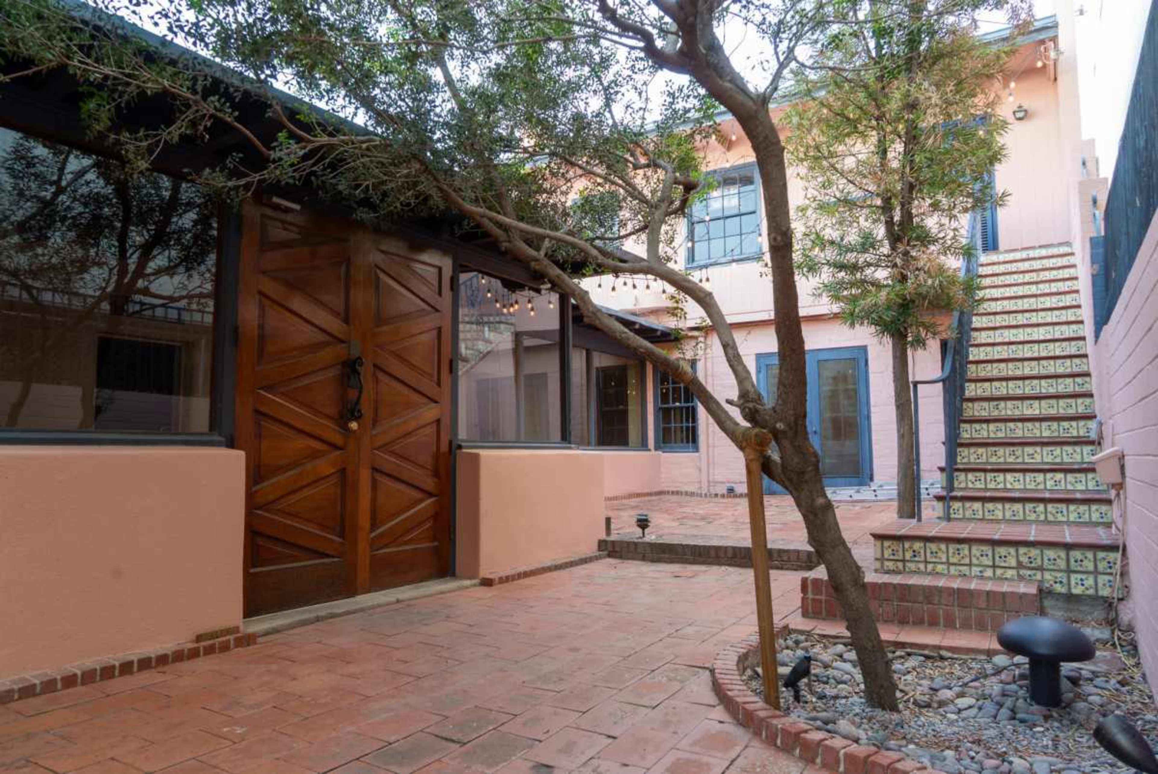 The image shows a courtyard with a wooden door, a tree, and a staircase leading to a building, all surrounded by brick and stone pathways.