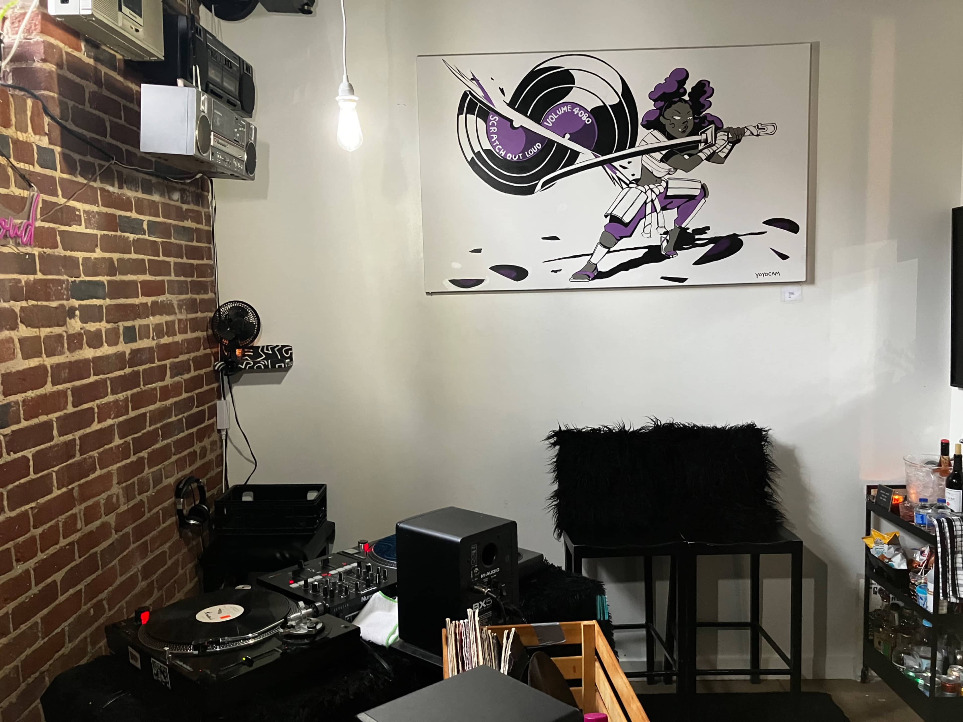 The room features a turntable setup on a black surface against a brick wall, with a large framed artwork of a musician playing an electric guitar above it.