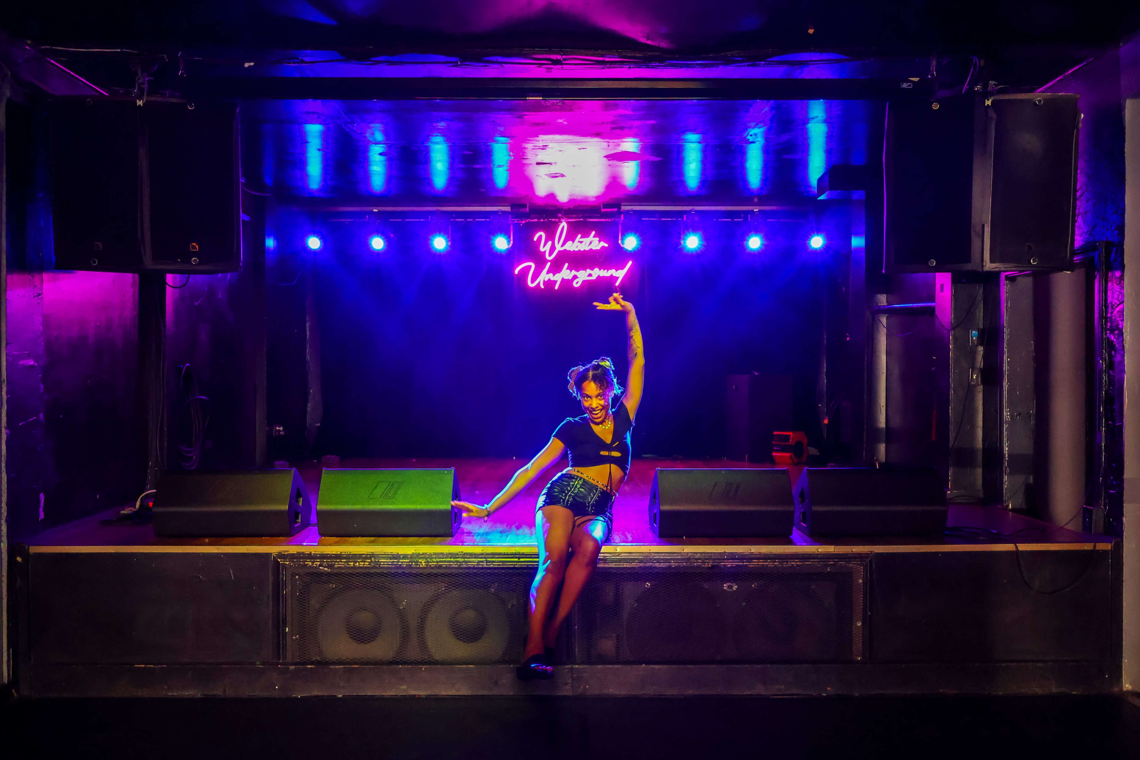 A performer poses on stage in front of vibrant purple and blue lighting with sound equipment set up.