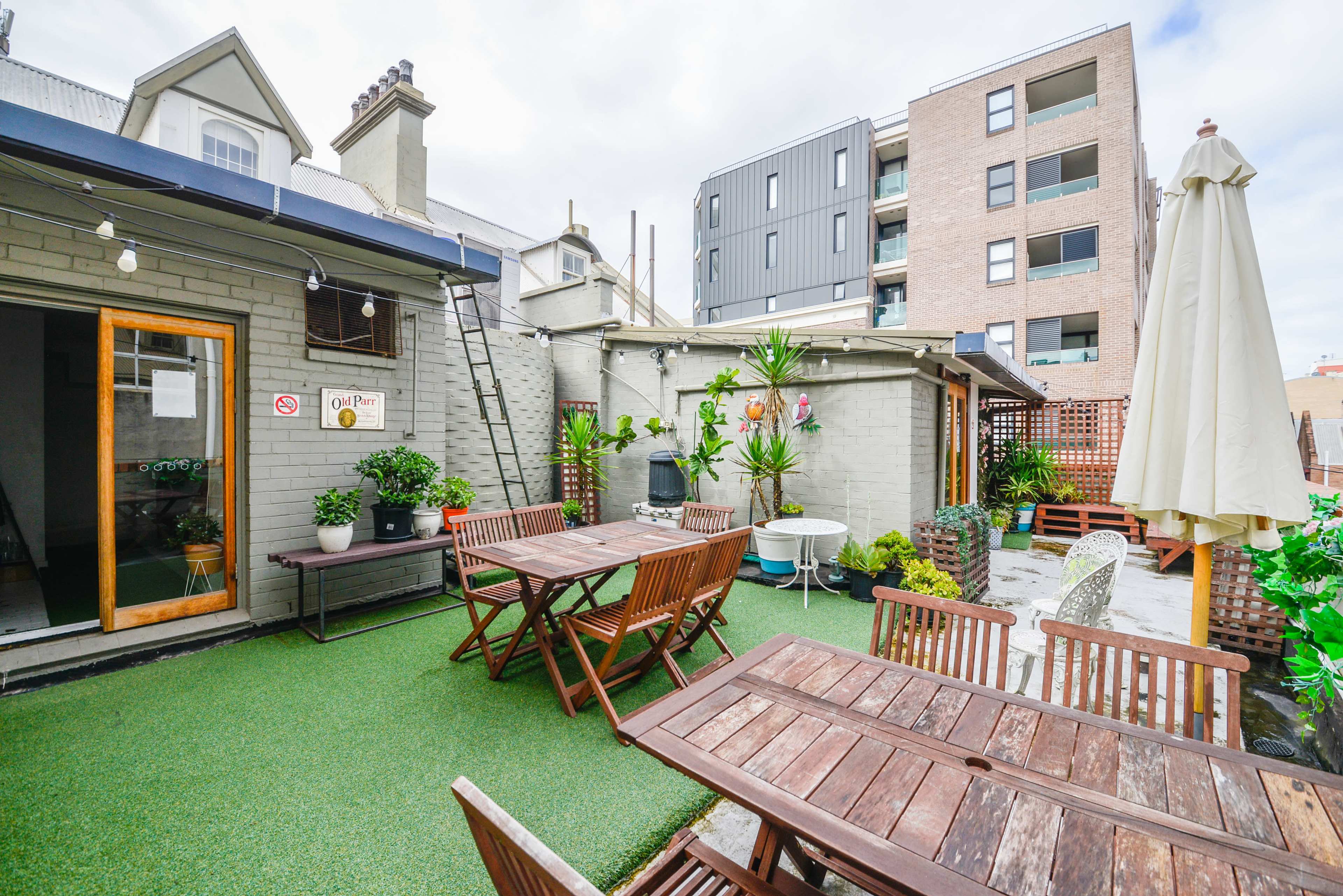 A patio area with wooden tables and chairs, surrounded by potted plants, against a backdrop of both a traditional and modern building.