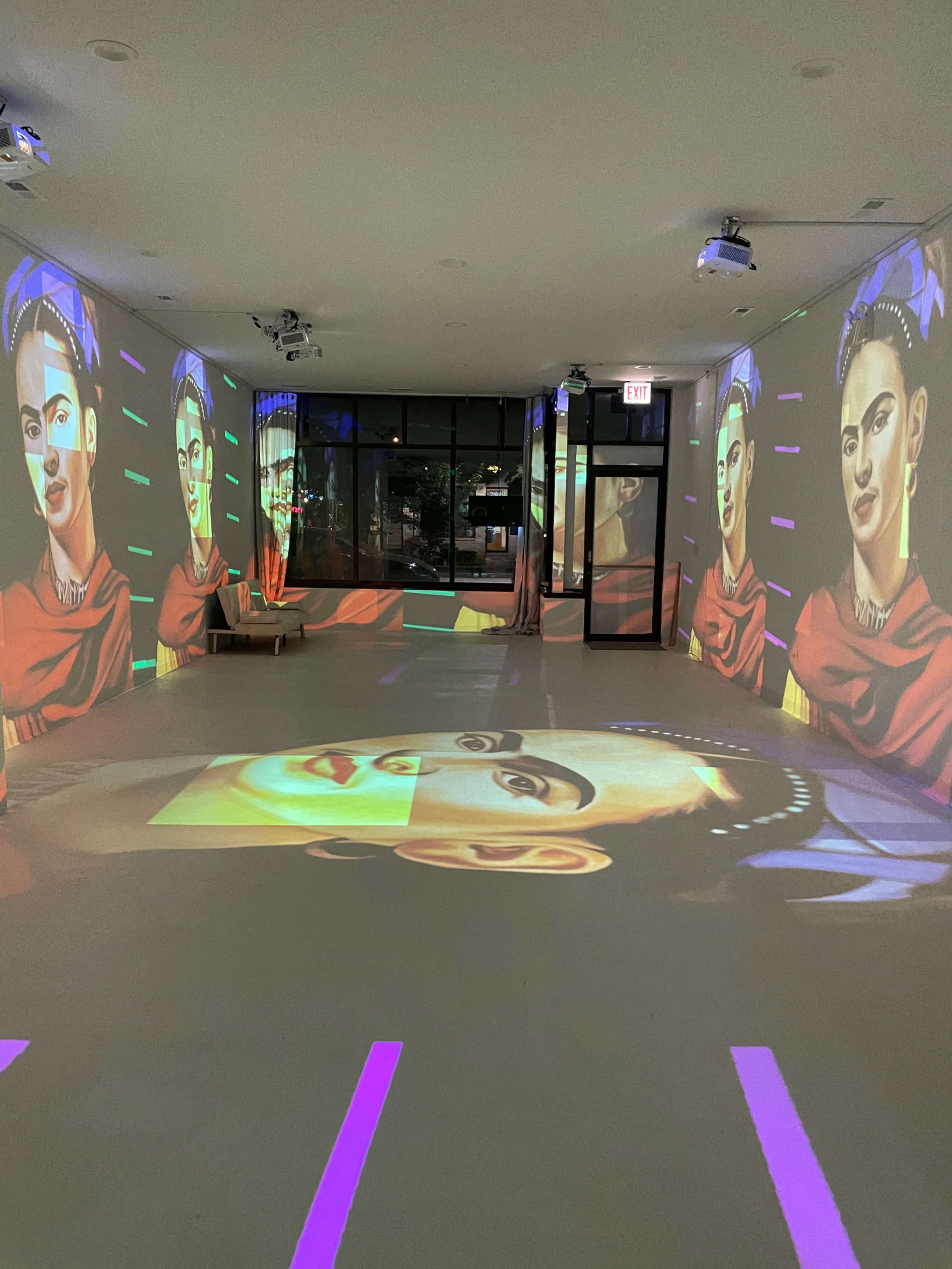 The room features large projected images of Frida Kahlo on the walls and floor, creating an immersive art installation.