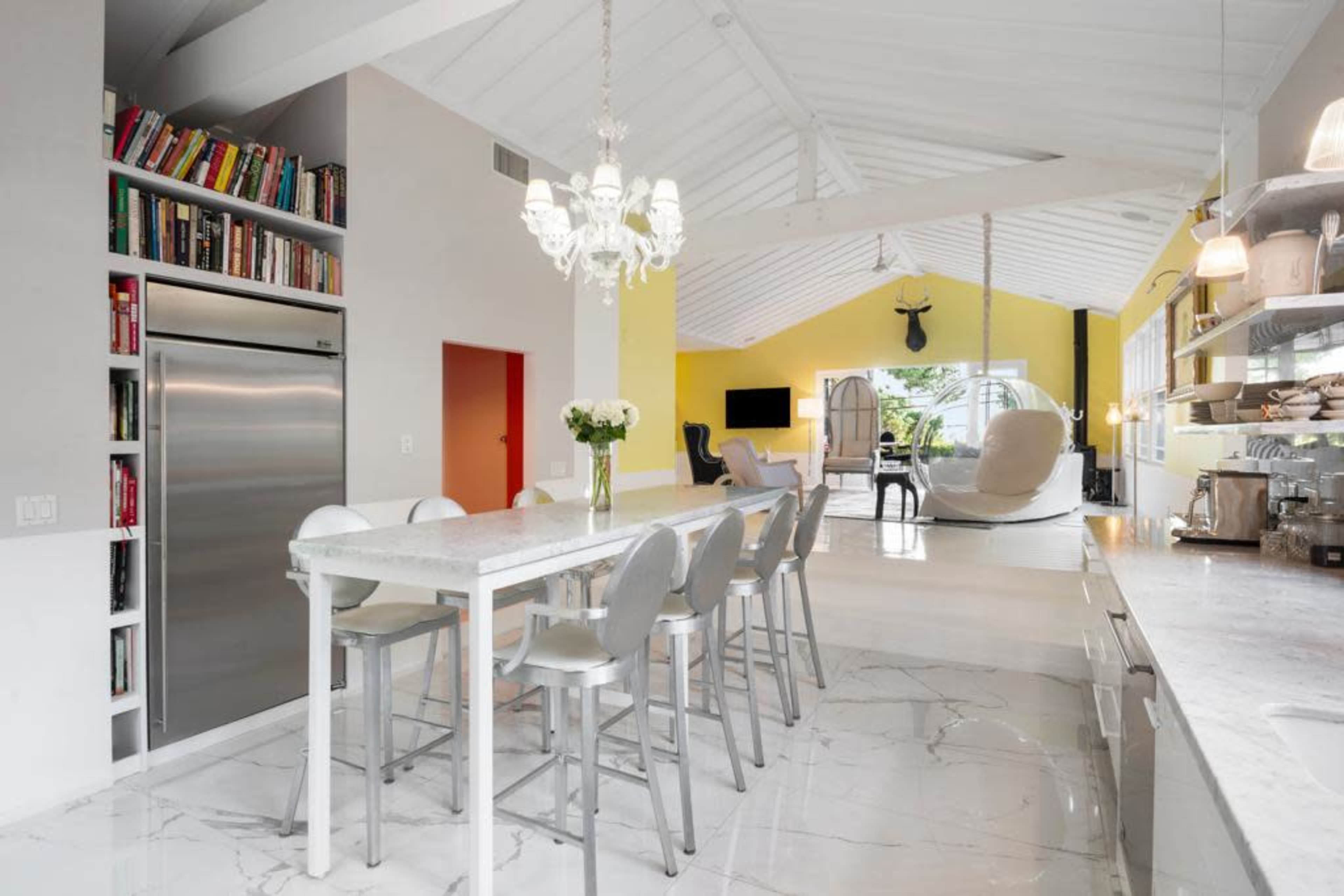 A modern kitchen features a long white dining table with high chairs, large windows, and an open layout leading to a bright yellow living area.