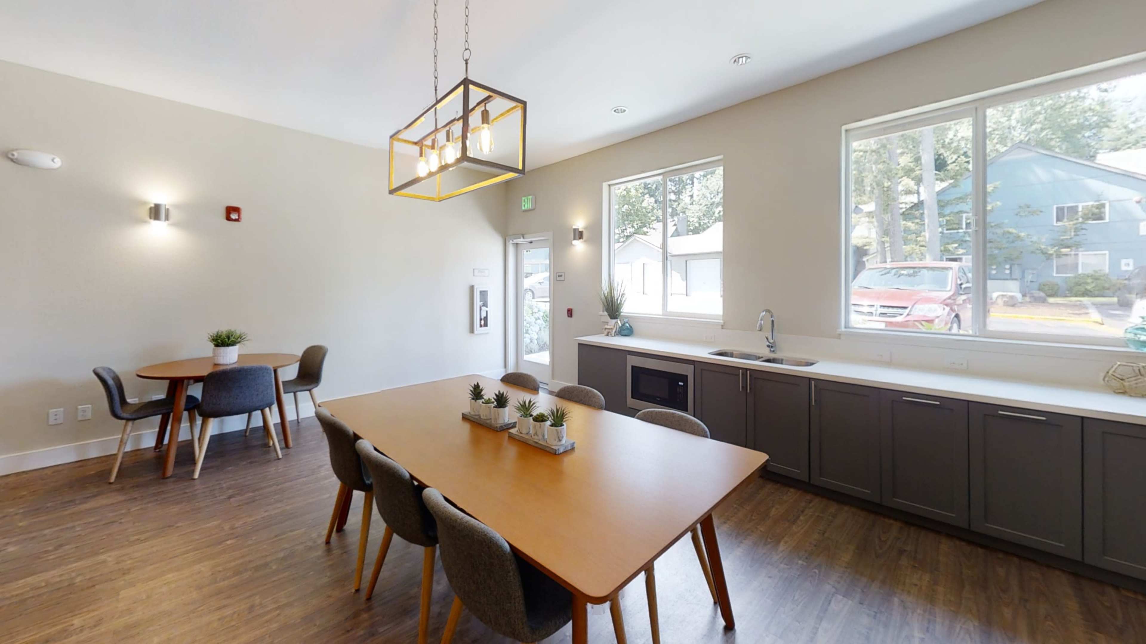 The image shows a spacious room featuring a large dining table with chairs, a smaller circular table, and large windows allowing natural light to enter.