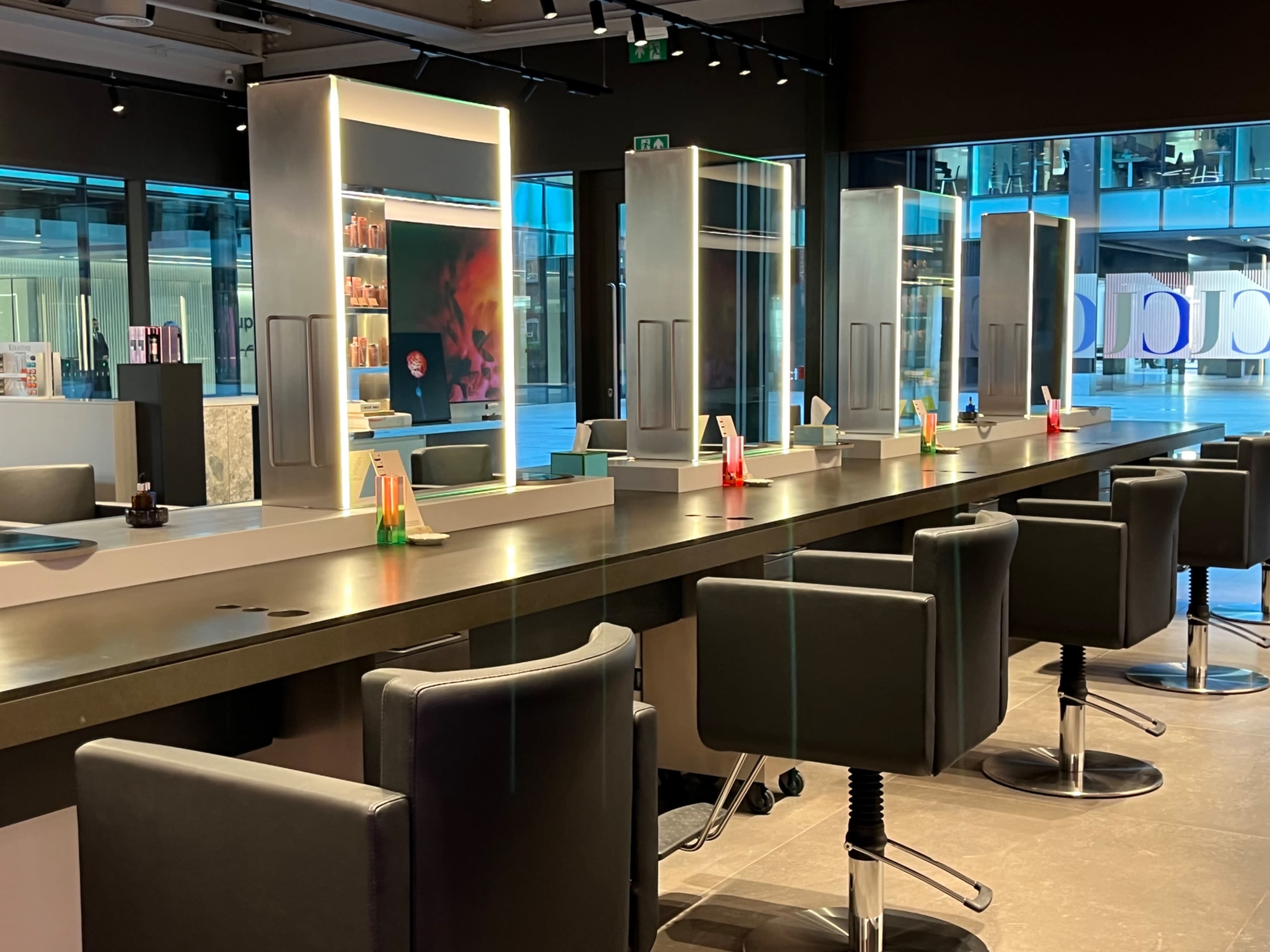 A hair salon features several black chairs and illuminated mirrors along a dark countertop, with colorful styling products displayed behind the mirrors.
