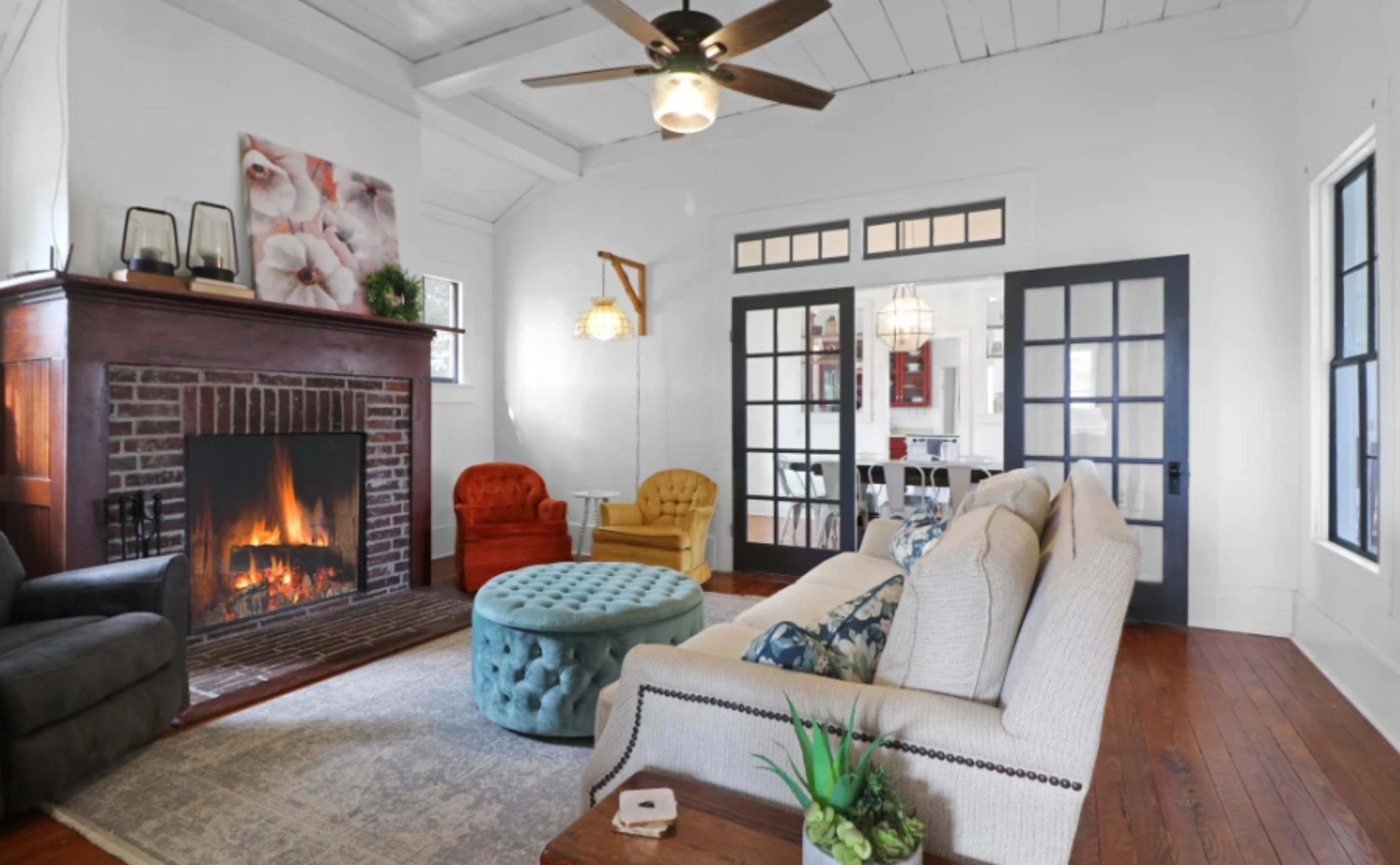 The living room features a fireplace, comfortable seating, and a blue ottoman, with large windows allowing natural light to enter.