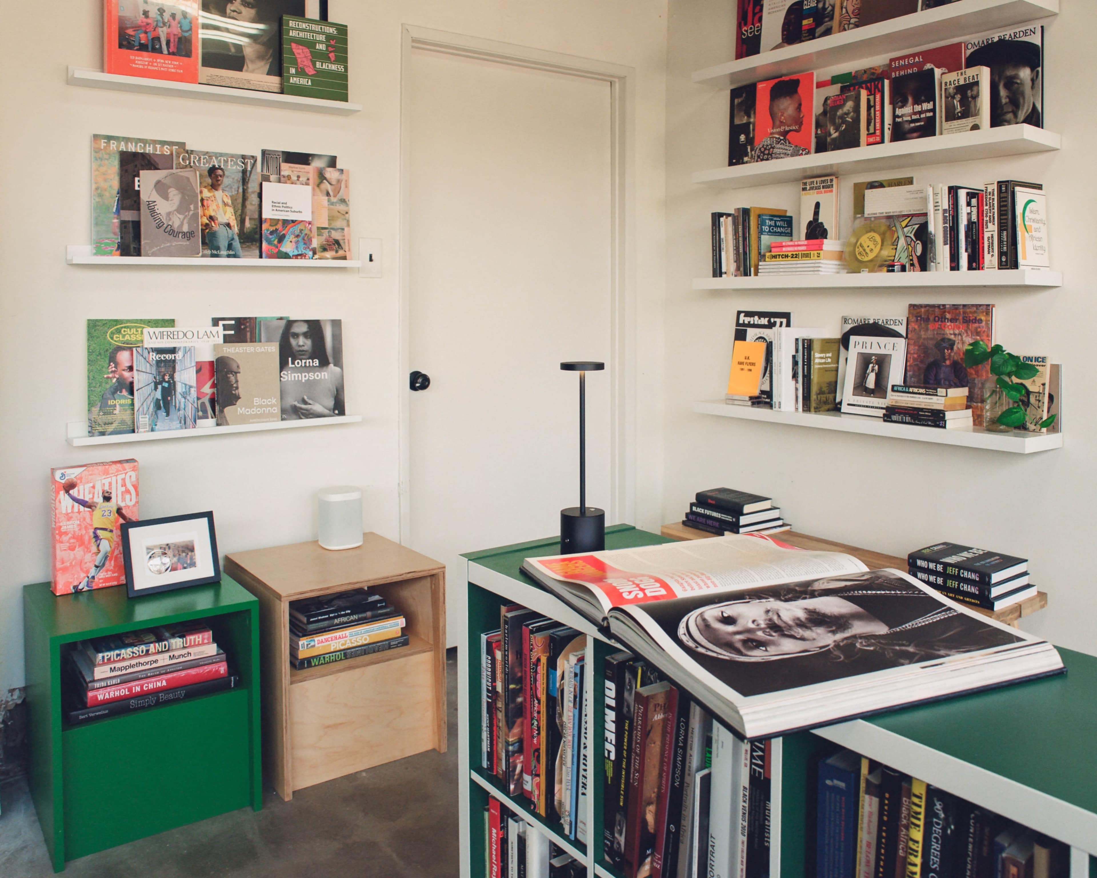 A cozy reading room features green furniture, shelves filled with various books and magazines, and a lamp on a table with an open magazine.