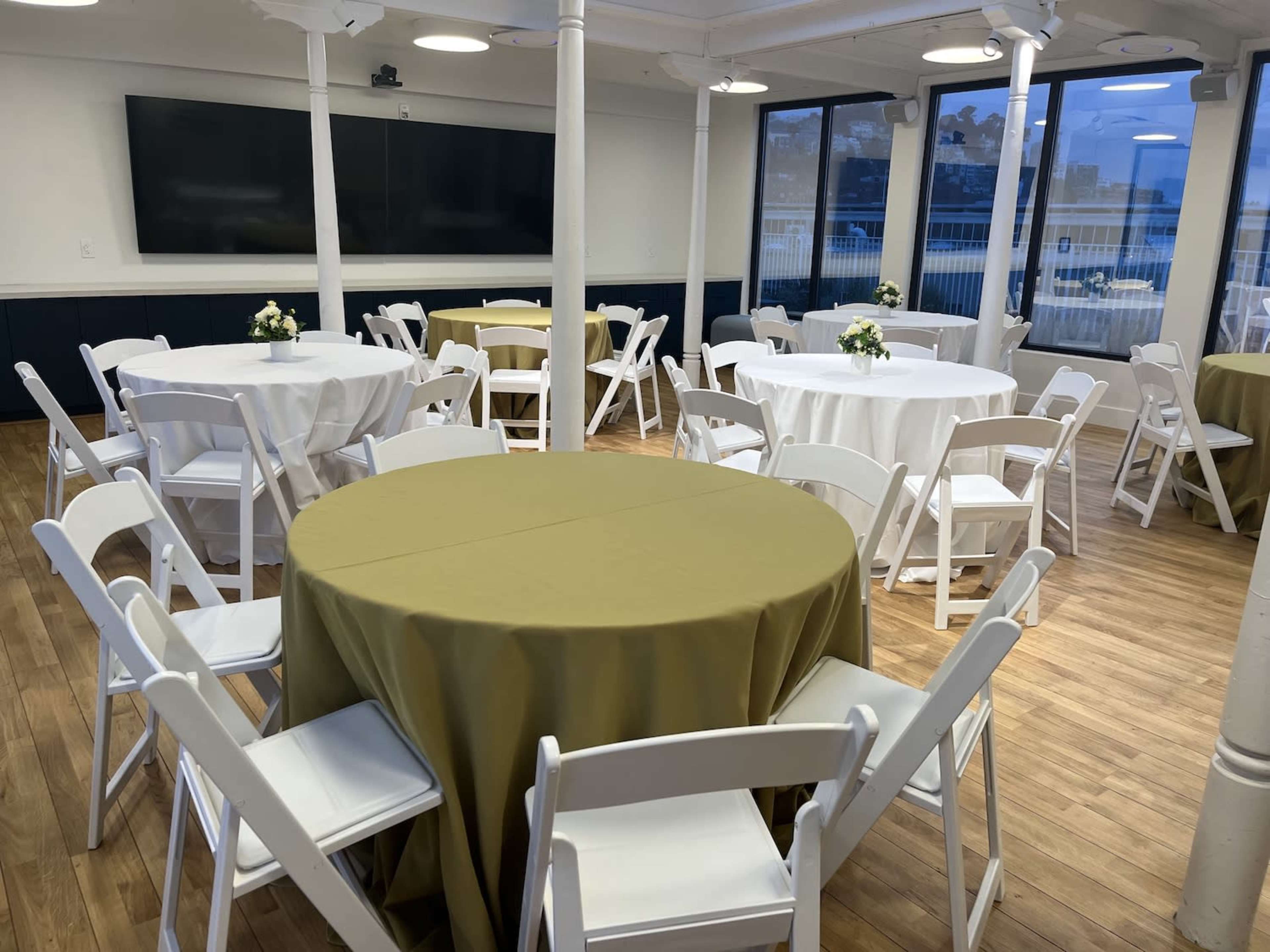 A room set up for an event, featuring several round tables with different colored tablecloths and white folding chairs arranged around them.