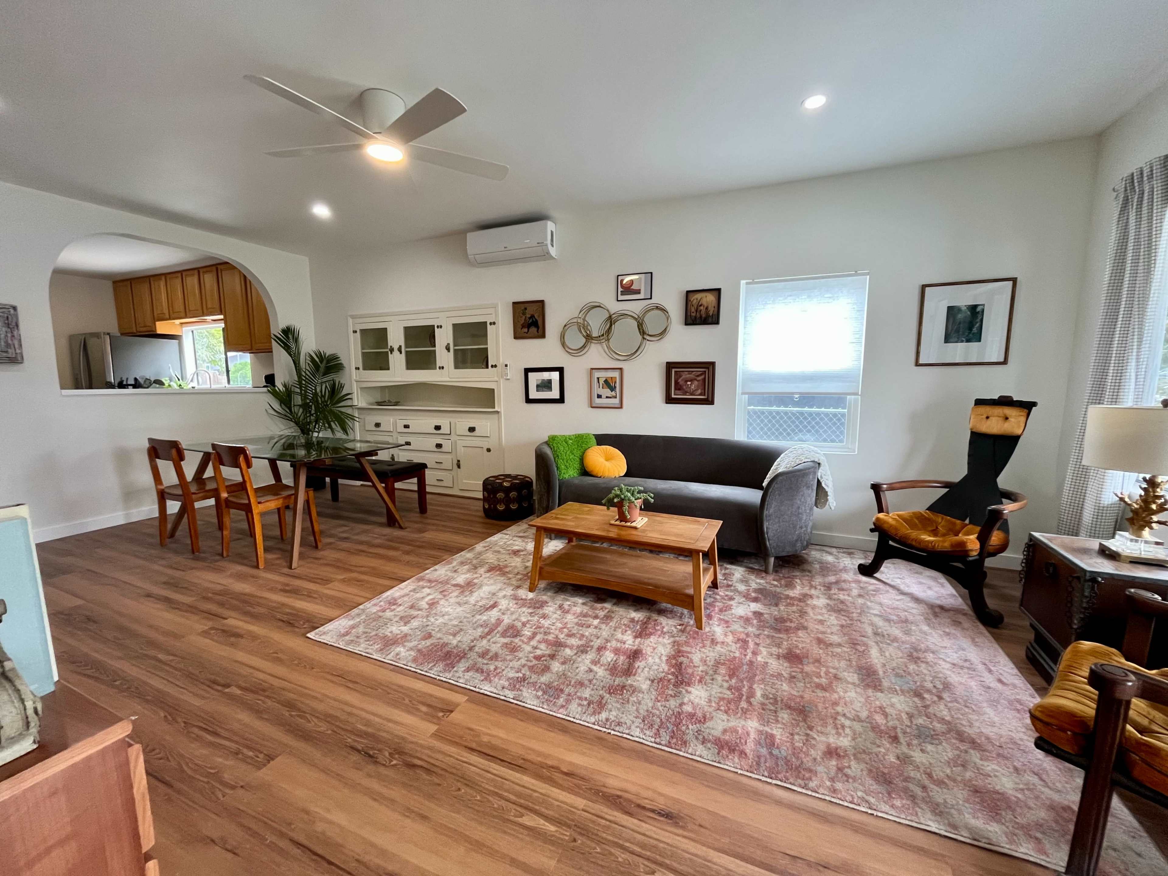 A cozy living room features a gray couch, wooden coffee table, and a dining area with chairs, surrounded by various wall art and décor.