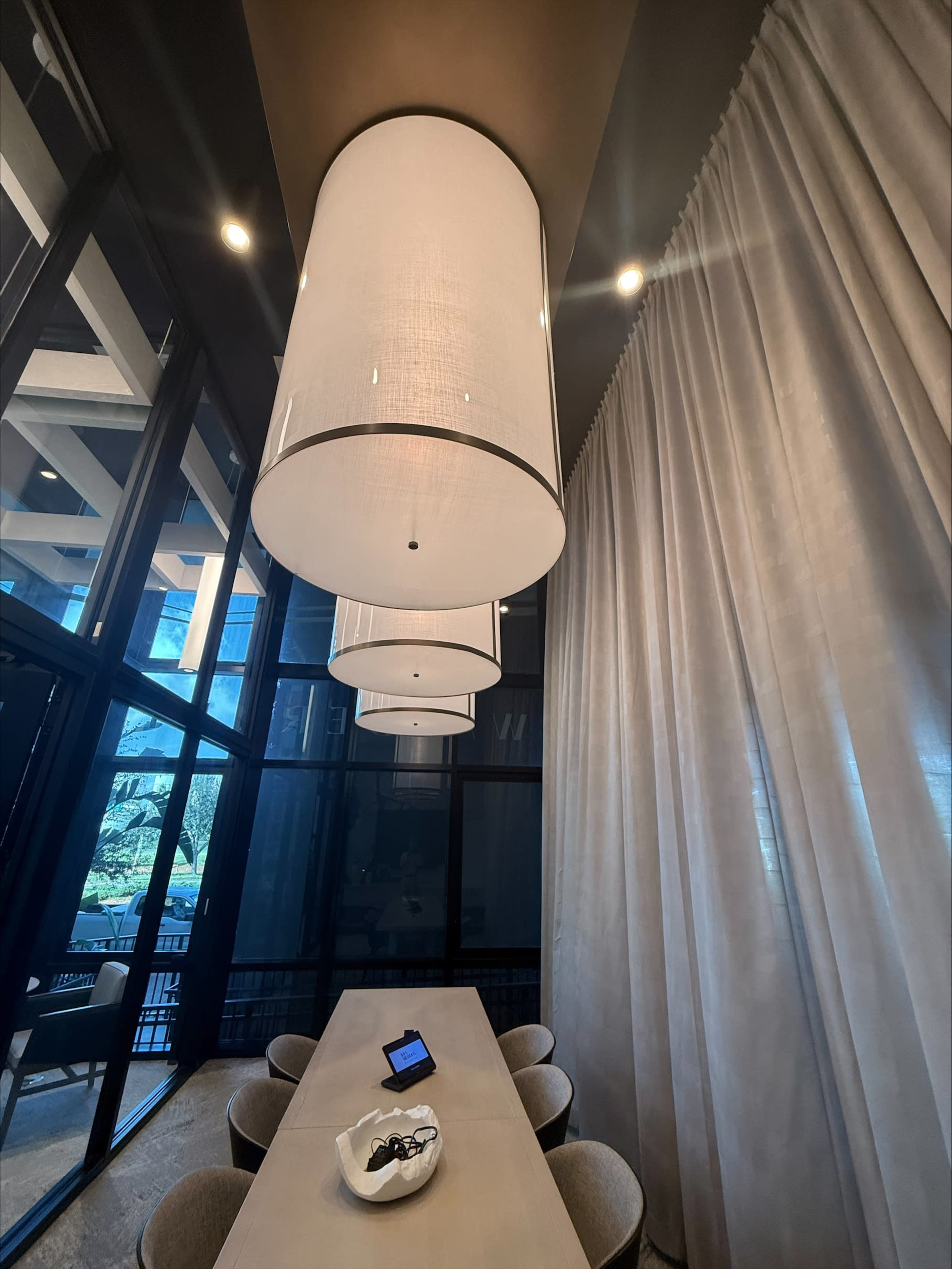 The image shows a modern dining area with a long table, surrounded by upholstered chairs, under three large pendant lights, and features curtains and large windows.