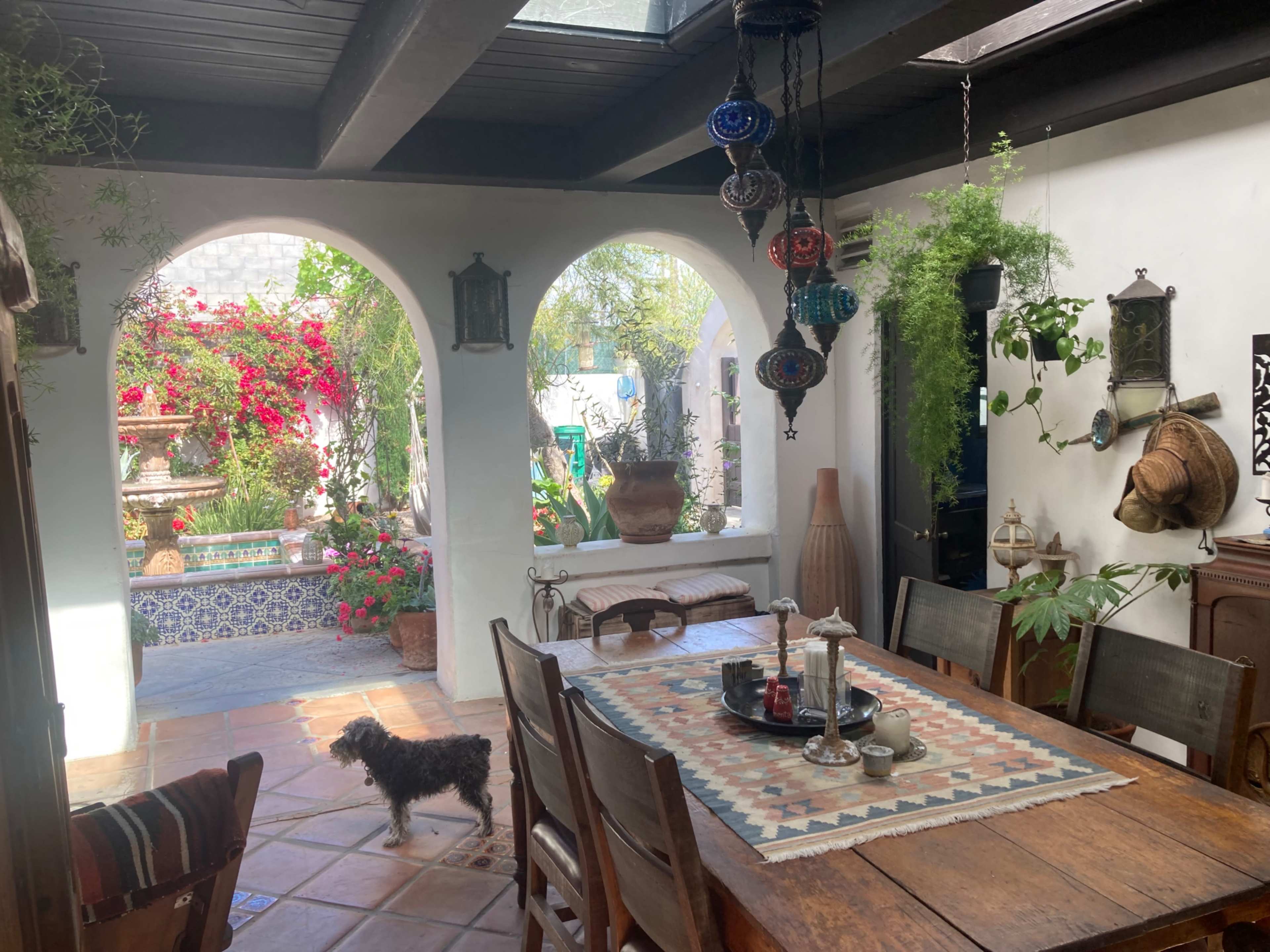 The image shows an indoor dining area with a wooden table and chairs, open arches leading to a garden filled with potted plants and colorful flowers.