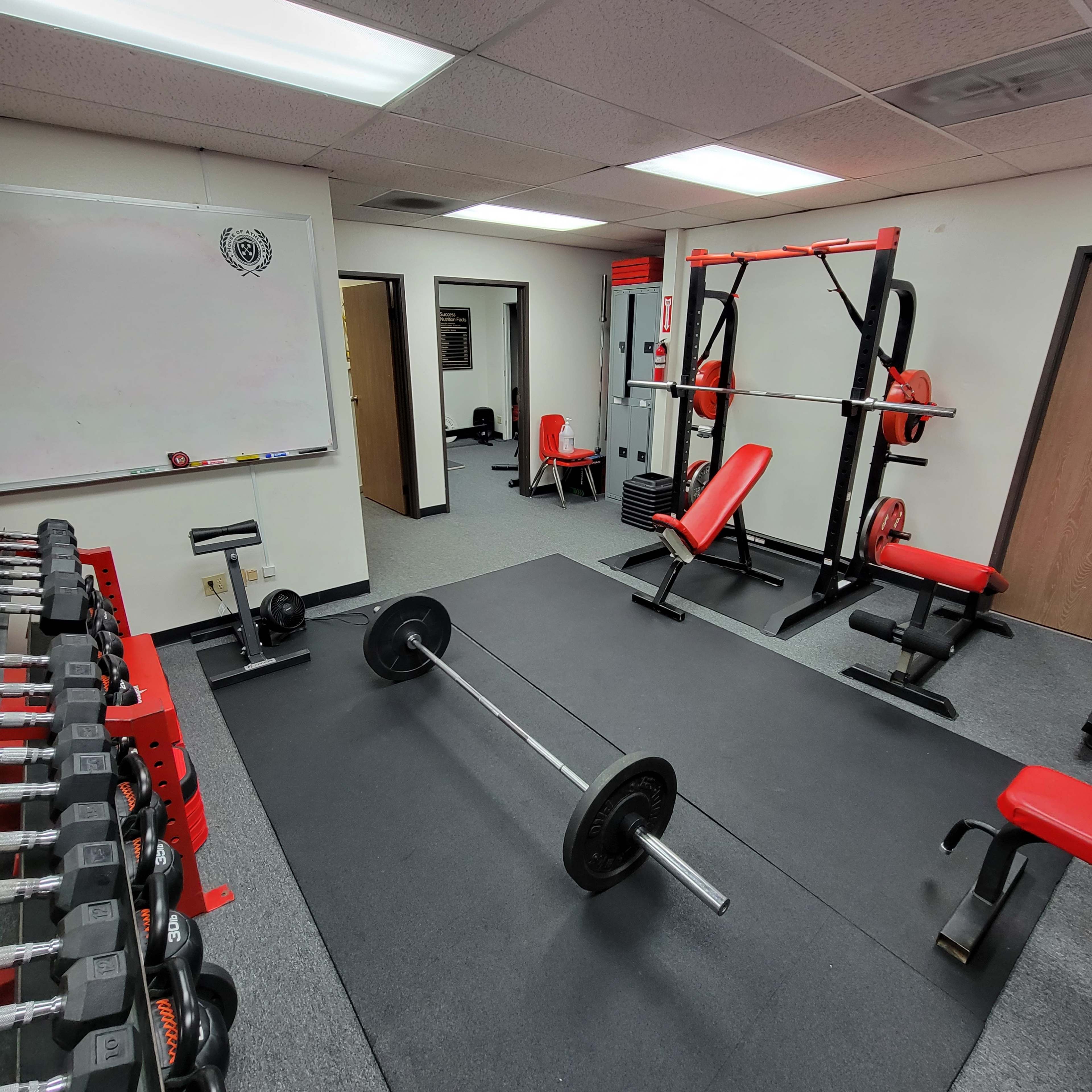 The gym features multiple weightlifting stations, including a barbell on the floor, dumbbell racks, and a squat rack with benches.