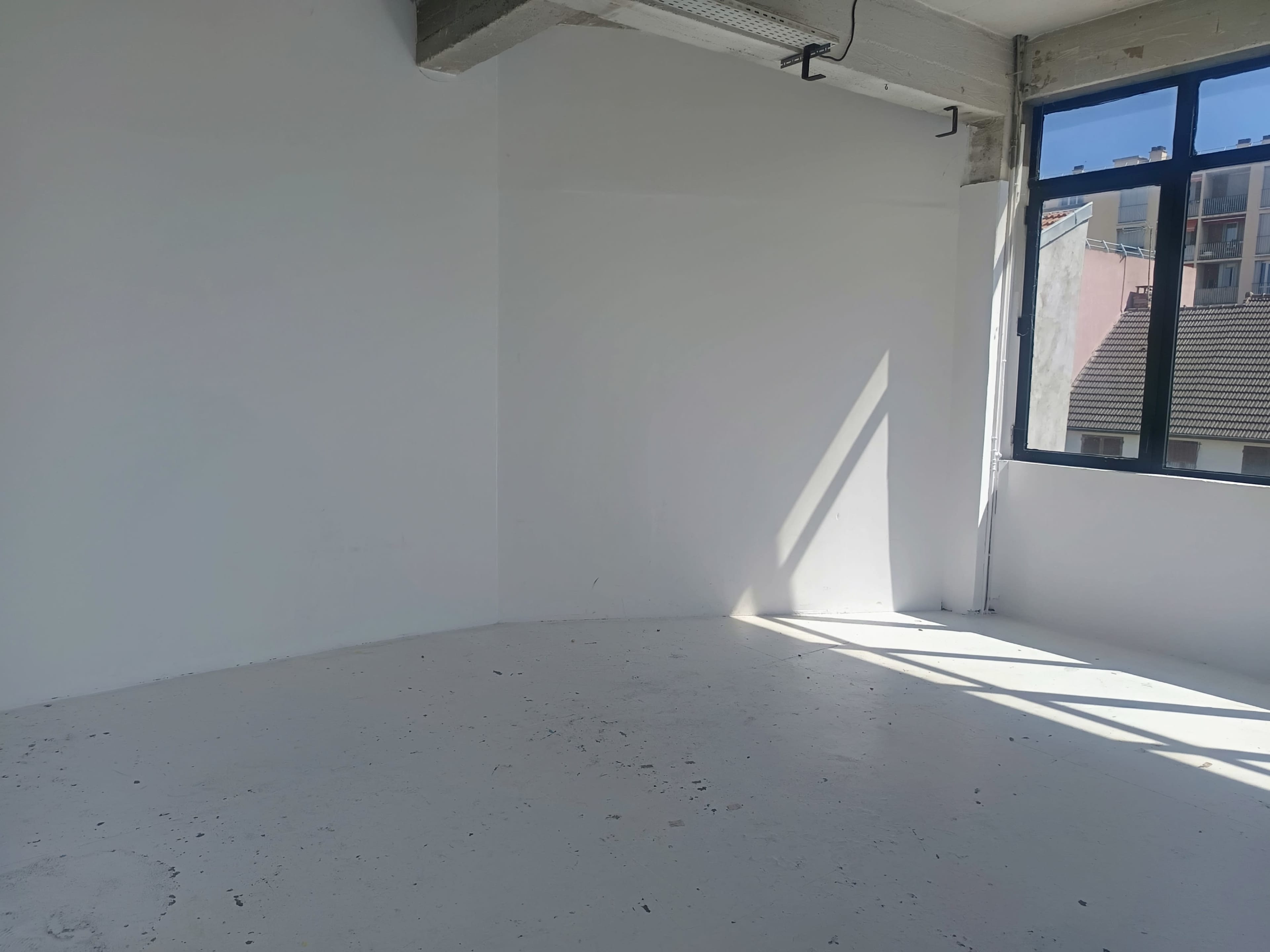 The image shows an empty room with a white wall and a large window allowing natural light to enter.