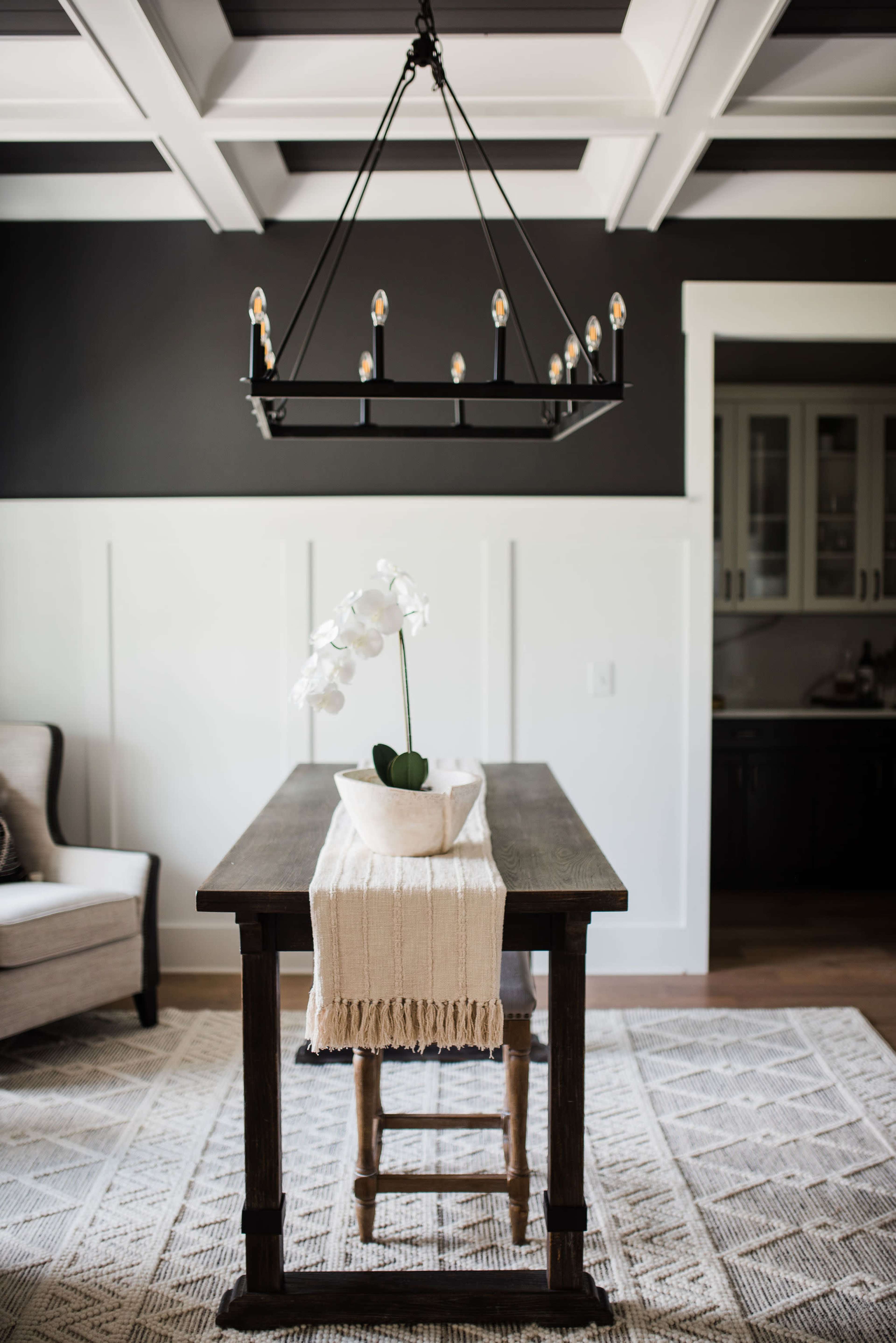 A wooden table with a white table runner and an orchid centerpiece is illuminated by a large black chandelier in a room with dark walls and coffered ceilings.