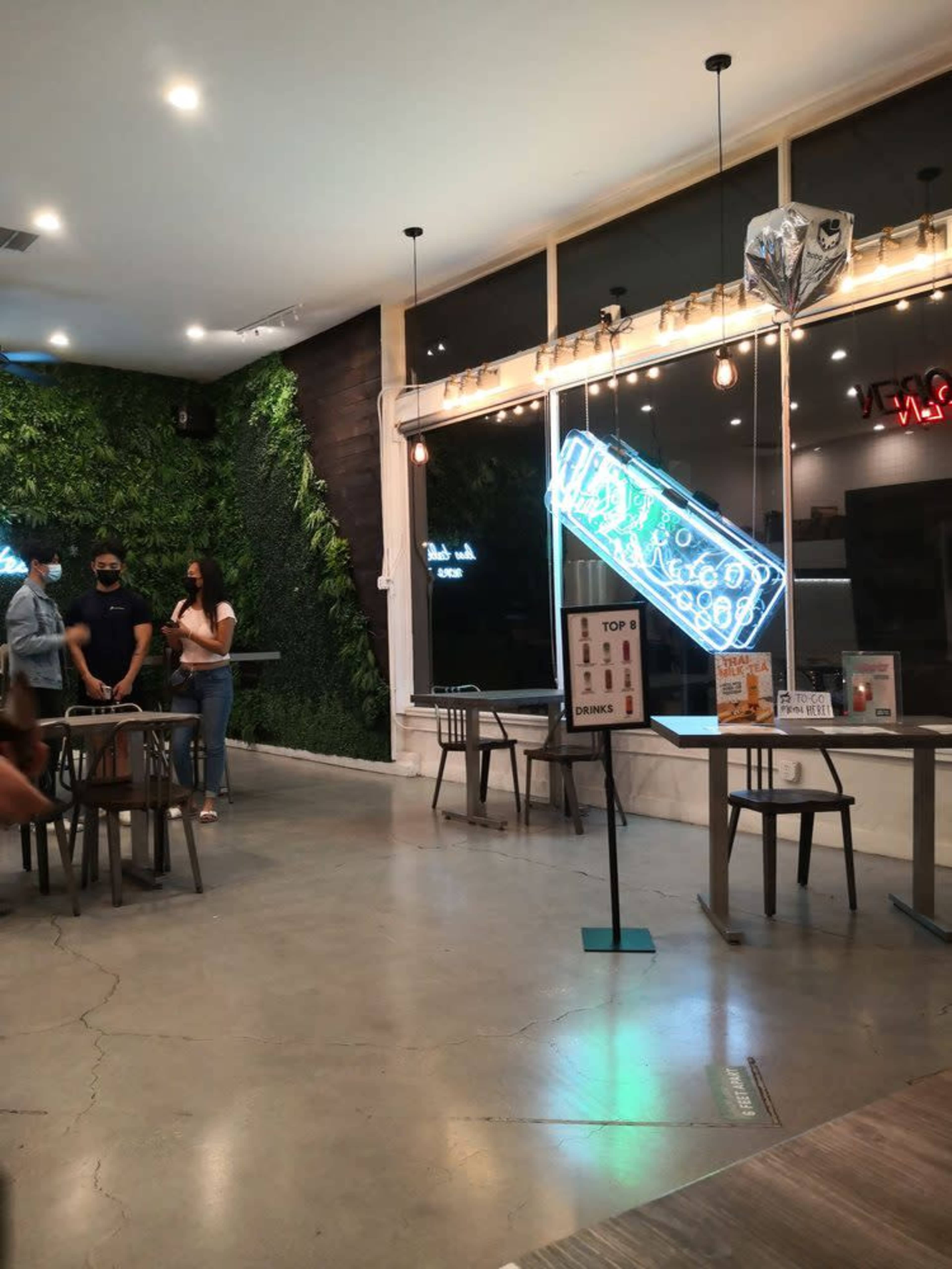 The image shows the interior of a cafe with green wall plants, a neon sign, and several tables and chairs arranged around the space.