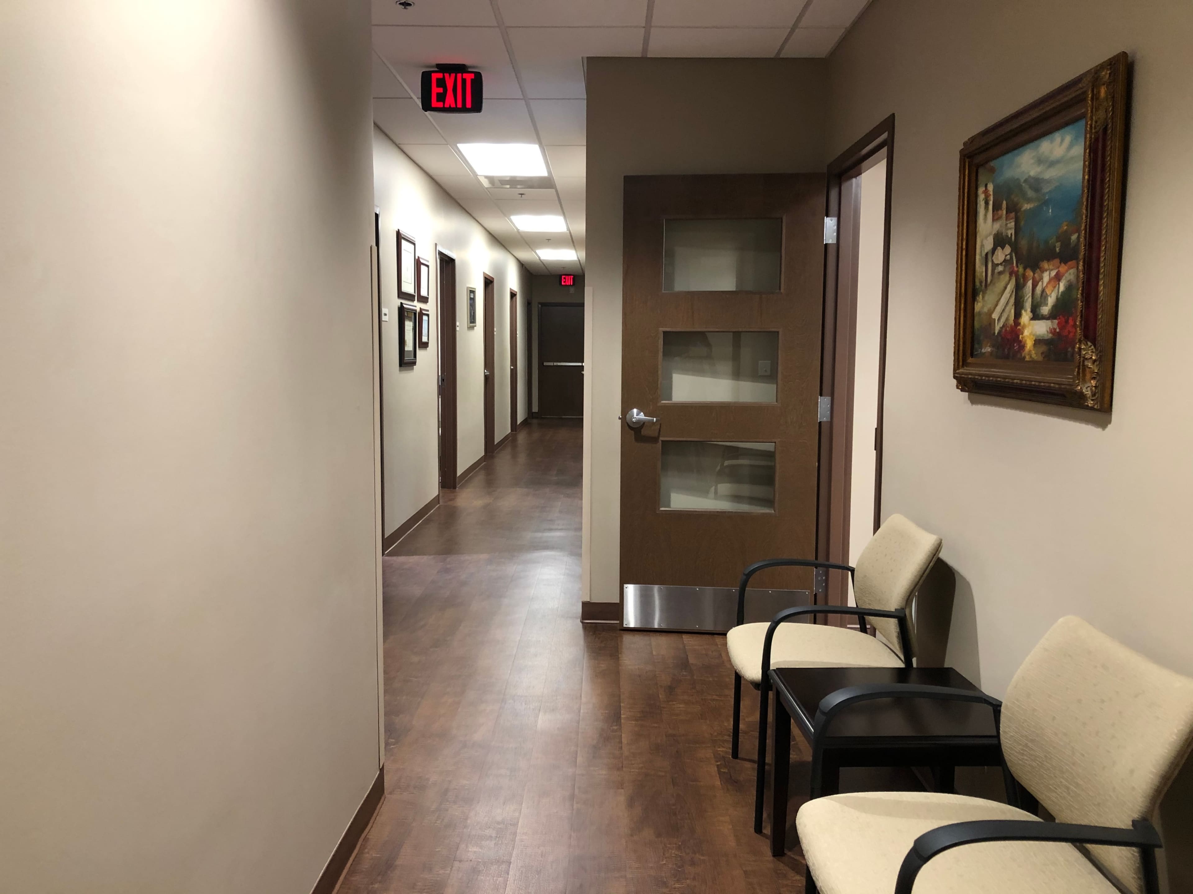 A well-lit corridor in a medical facility with beige walls, wooden flooring, and chairs arranged along one side.