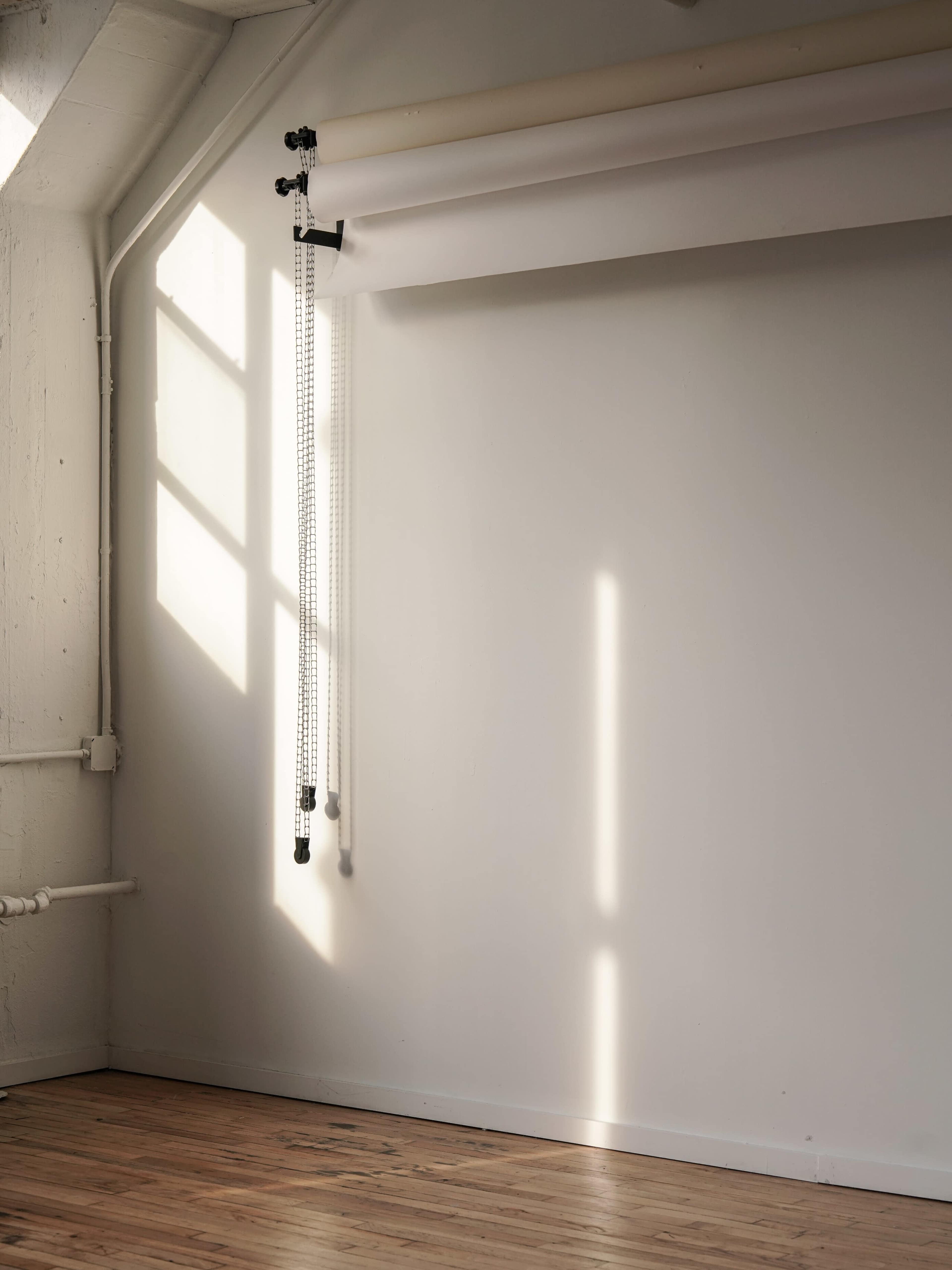 A minimalist interior space with a large window casting diagonal light onto a plain wall, alongside hanging rolls of paper.