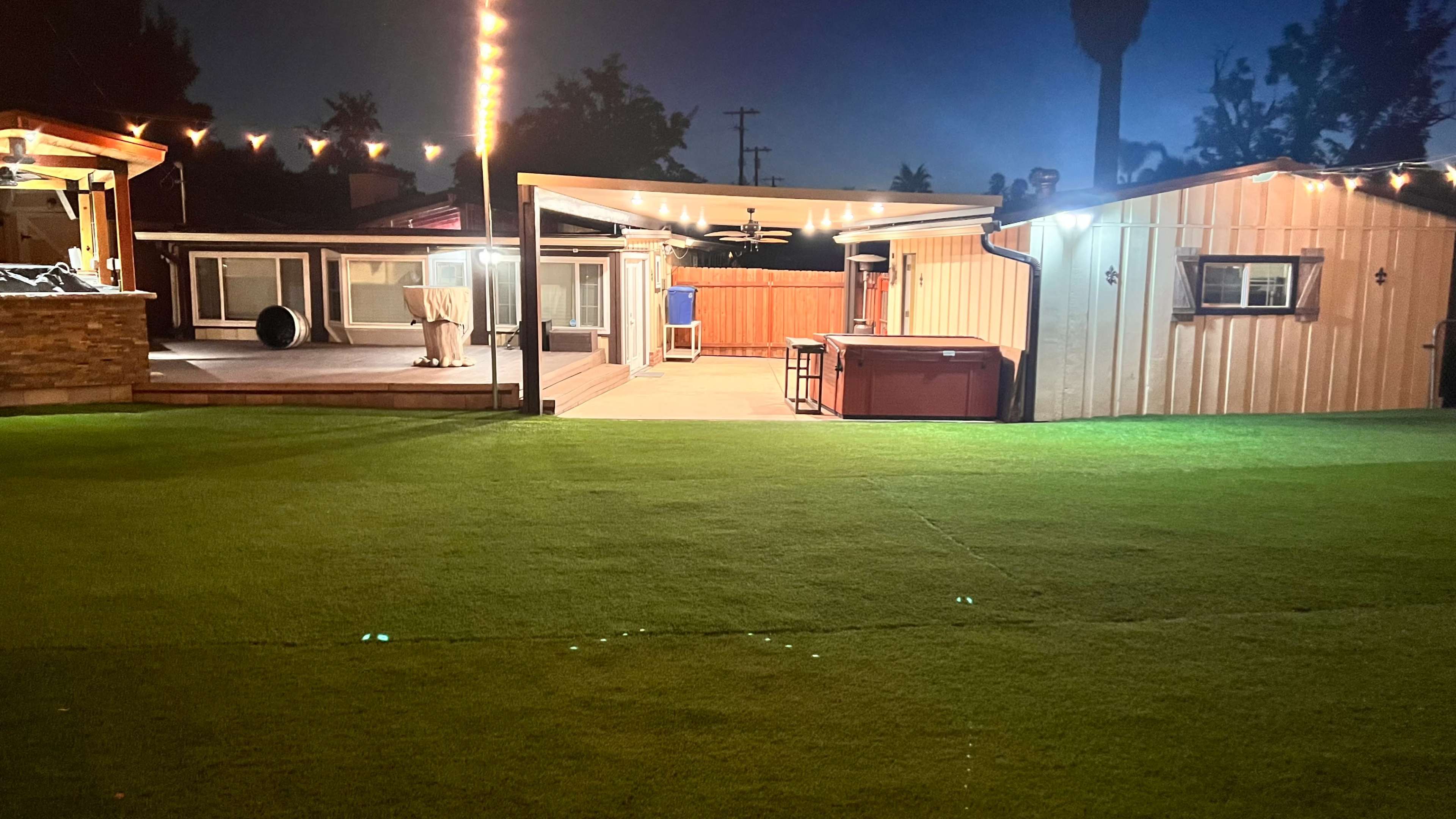 A backyard at night featuring a well-lit area with artificial grass, a patio with a bar, and a nearby shed.