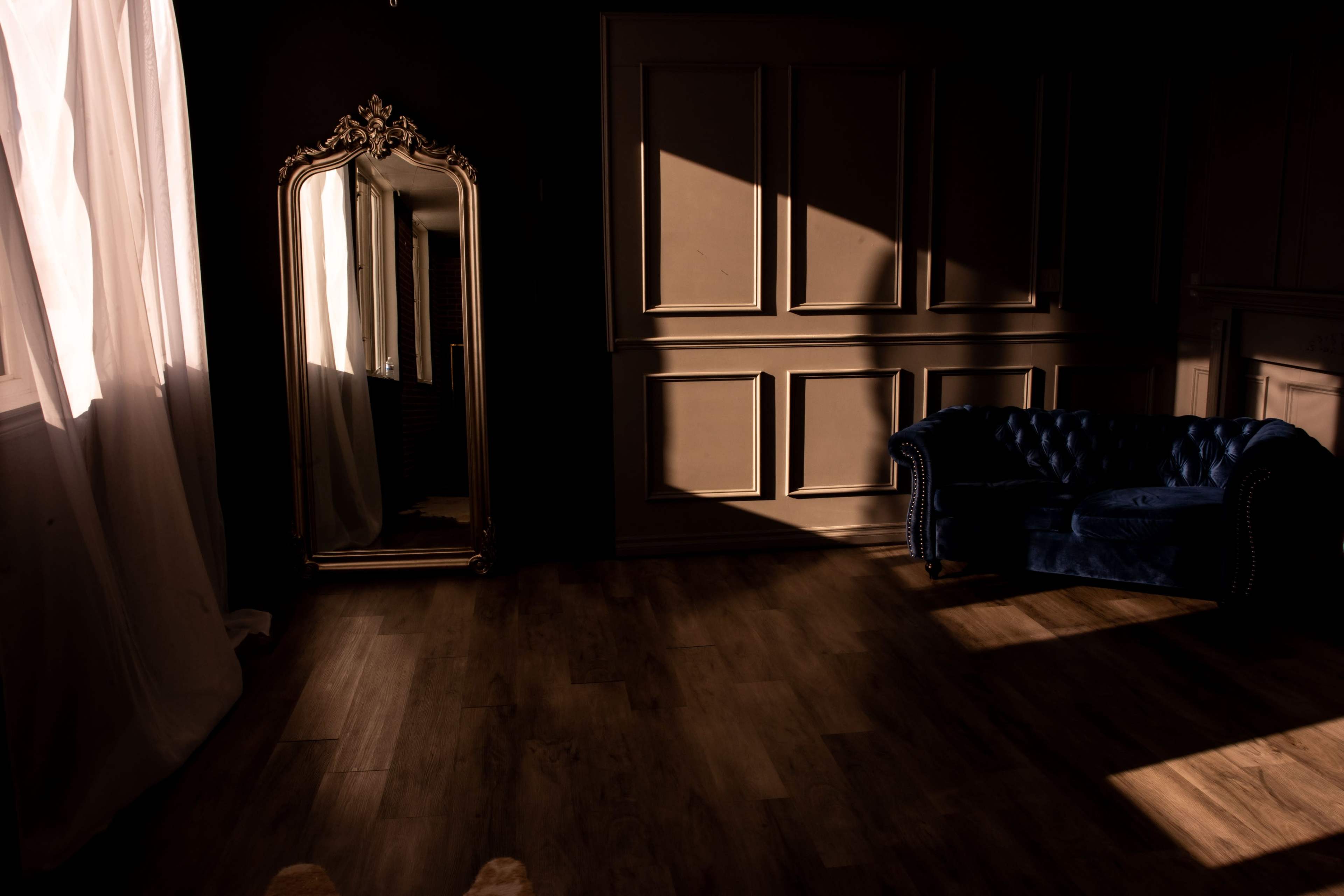 A dark room features a blue velvet couch and a decorative mirror reflecting light from a nearby window.