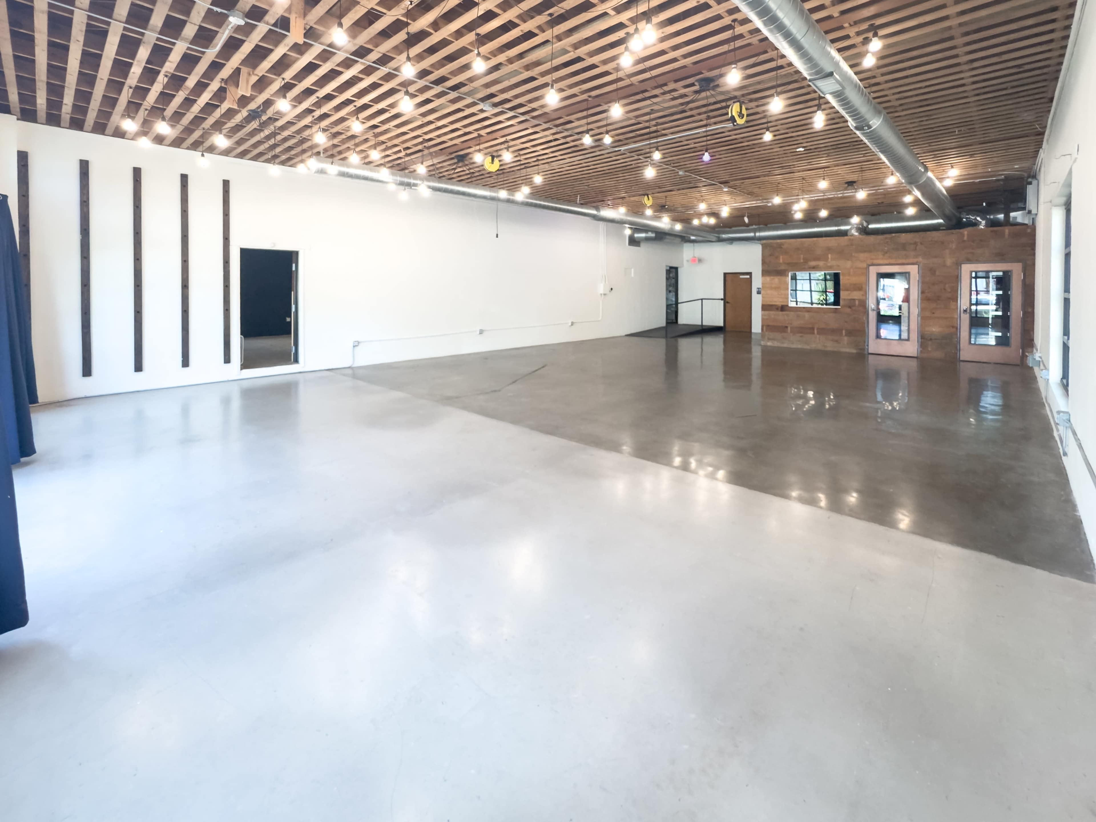 The image shows a spacious, empty interior room with polished concrete flooring and exposed wooden beams in the ceiling, illuminated by overhead lights.