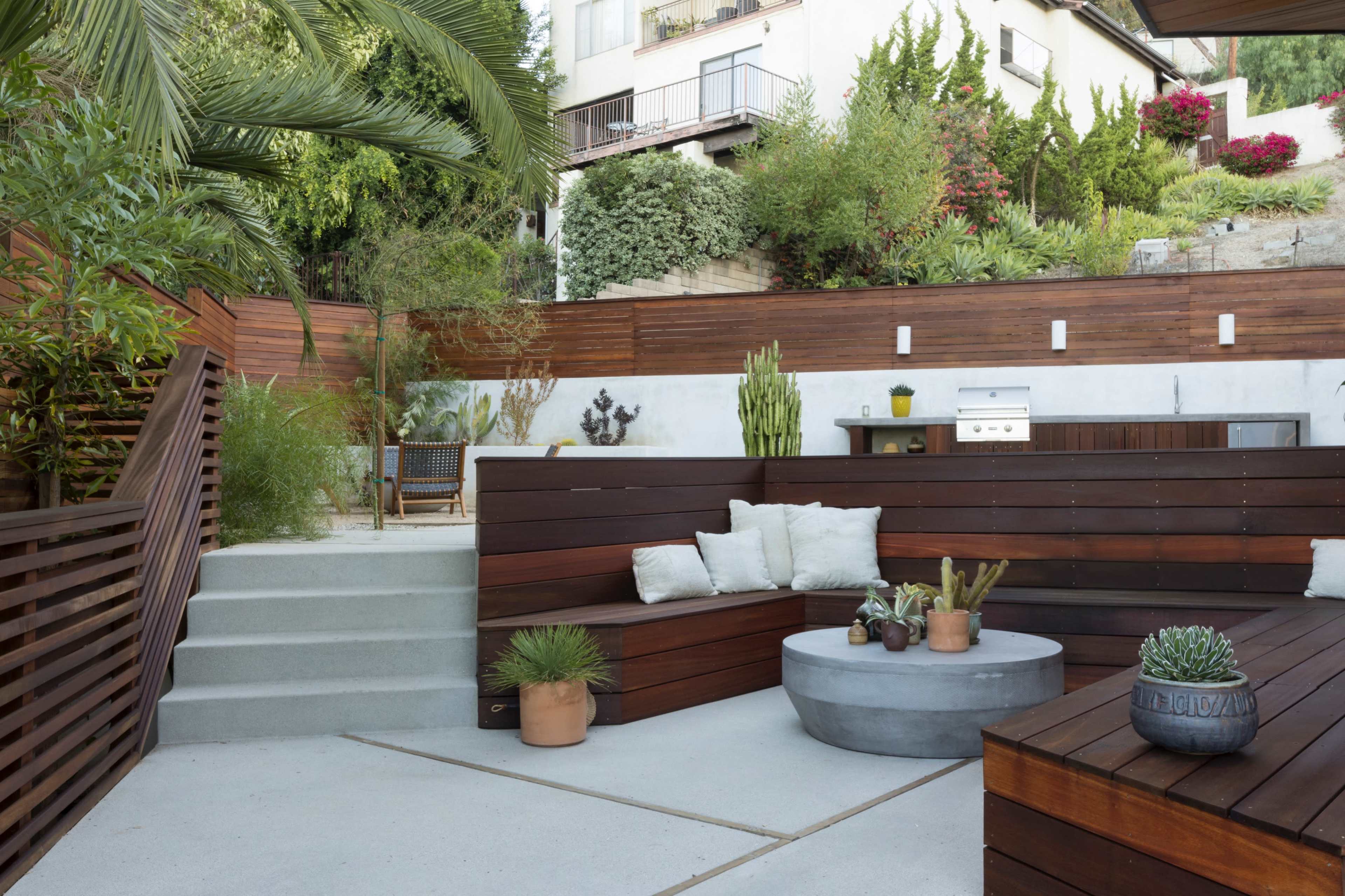 A modern outdoor seating area with wooden benches, a circular concrete table, and various plants, set against a backdrop of greenery and a hillside.