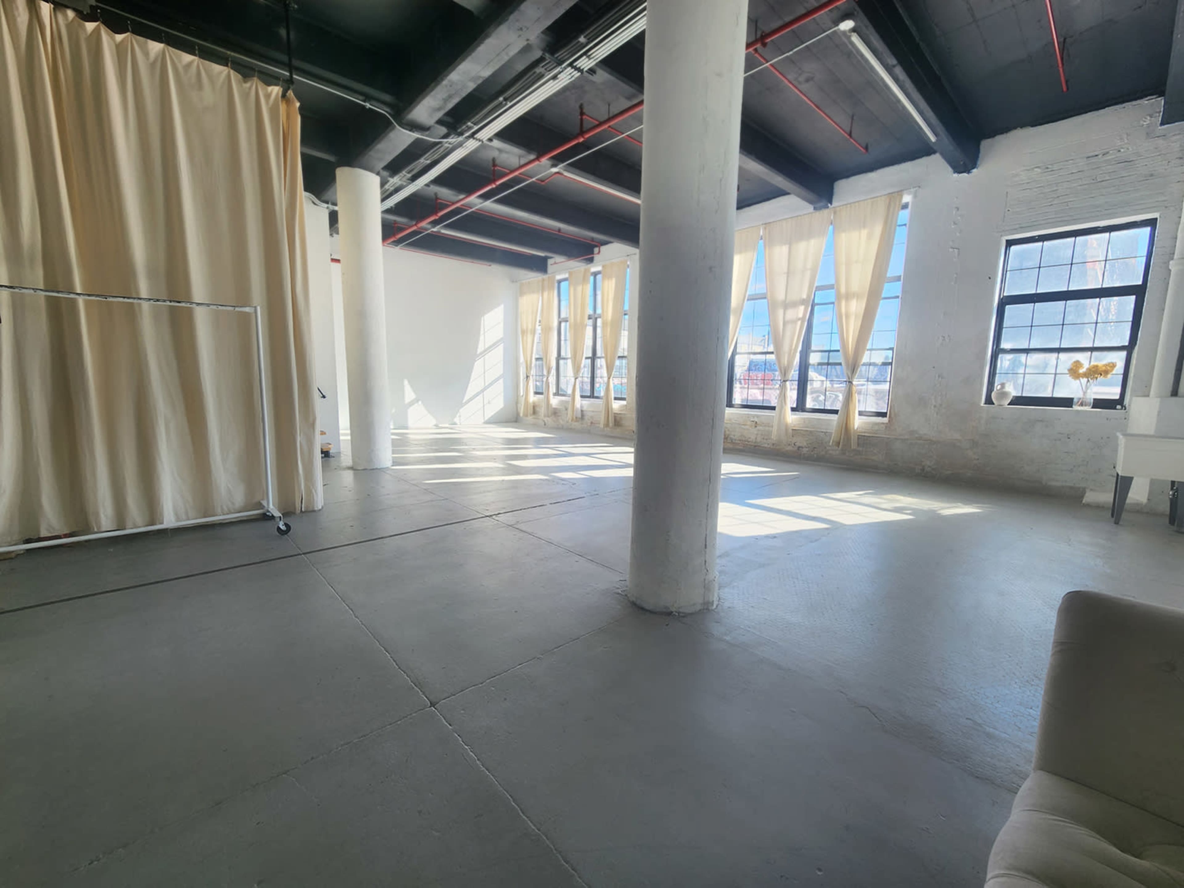 A spacious, well-lit room features tall windows with sheer curtains and exposed concrete pillars.