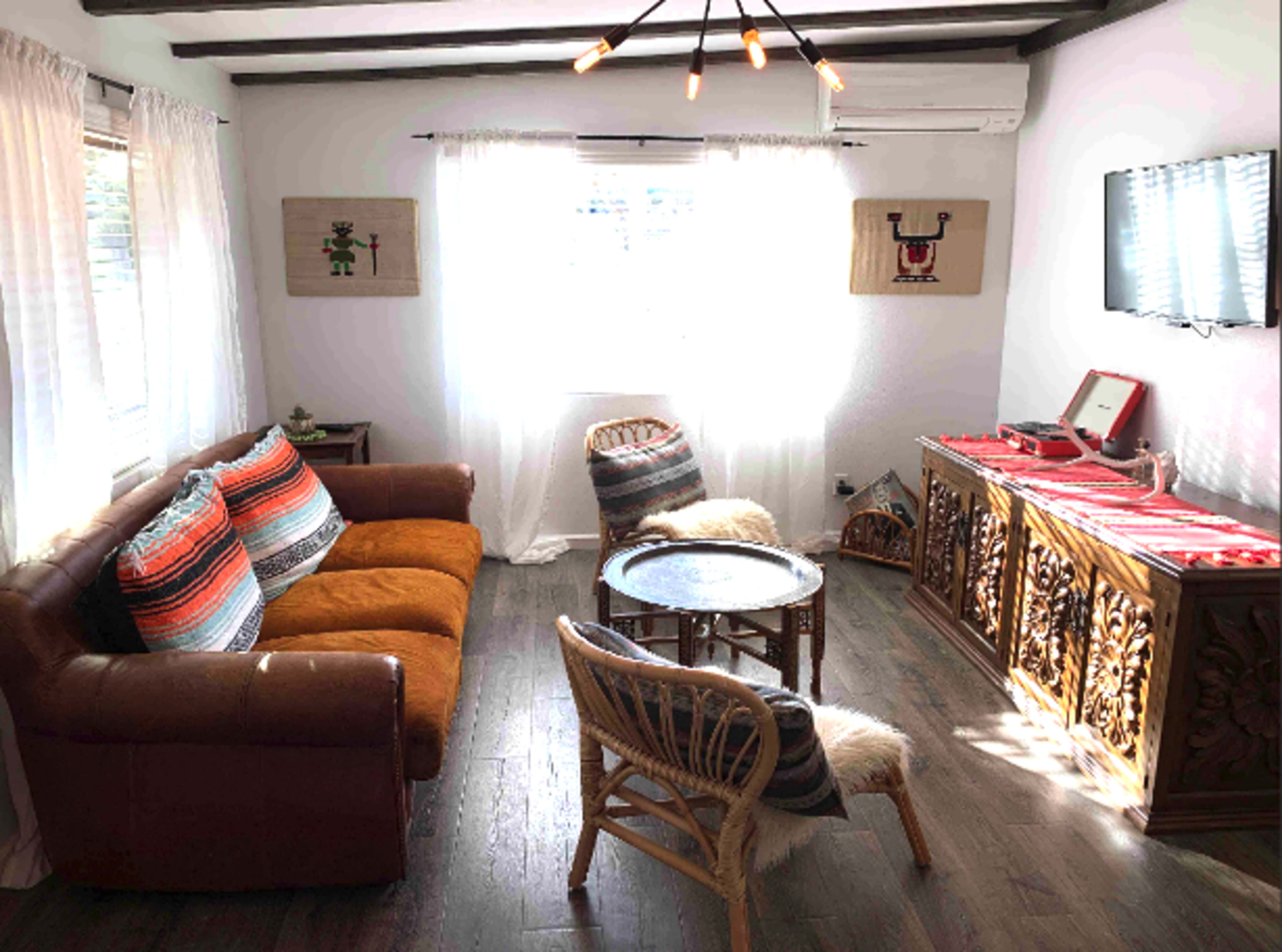 The image shows a cozy living room with a brown leather sofa, a round coffee table, and a rattan chair, illuminated by natural light coming through sheer white curtains.