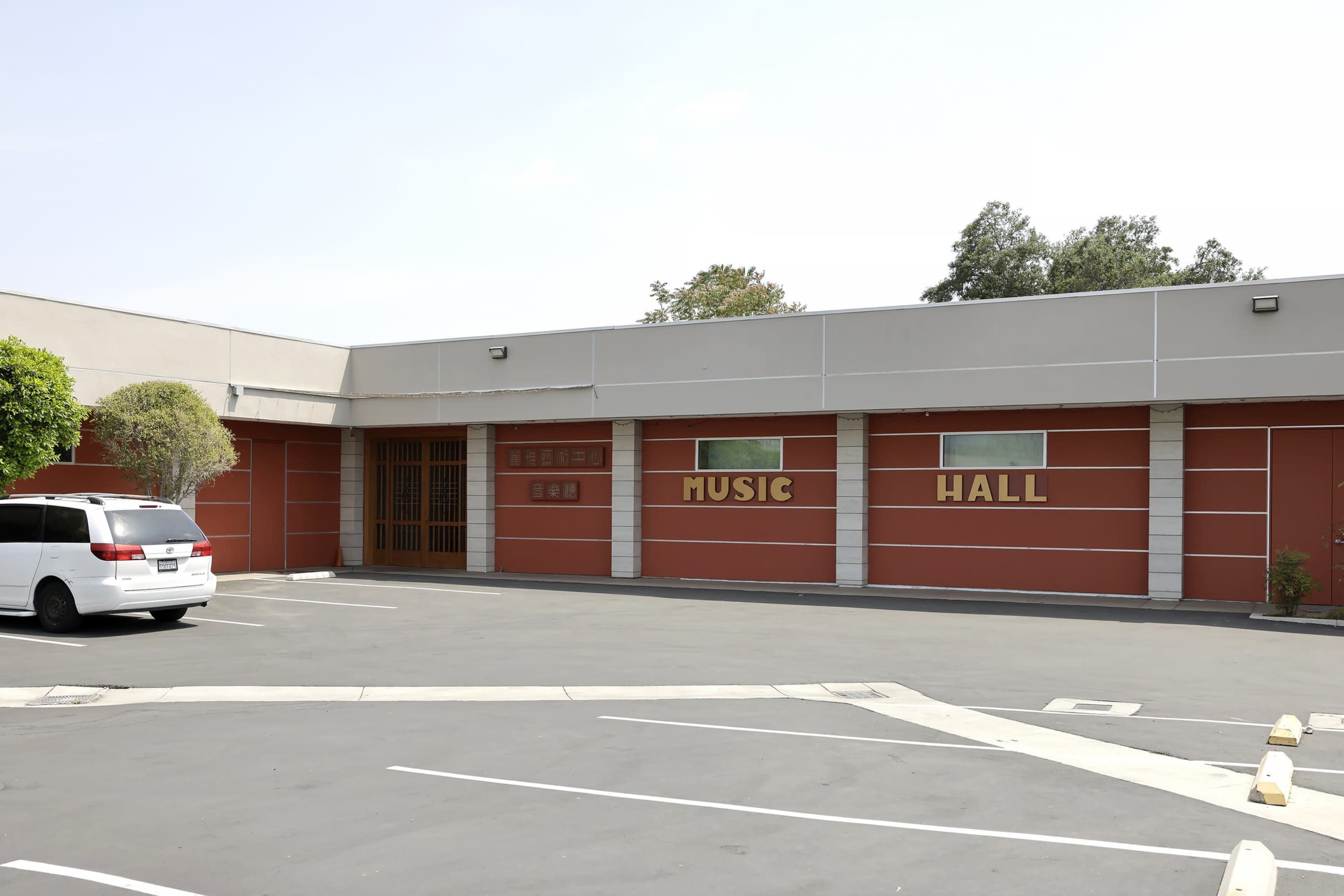 A red-brick building labeled "MUSIC HALL" with a parking lot in front and a white vehicle parked on the side.