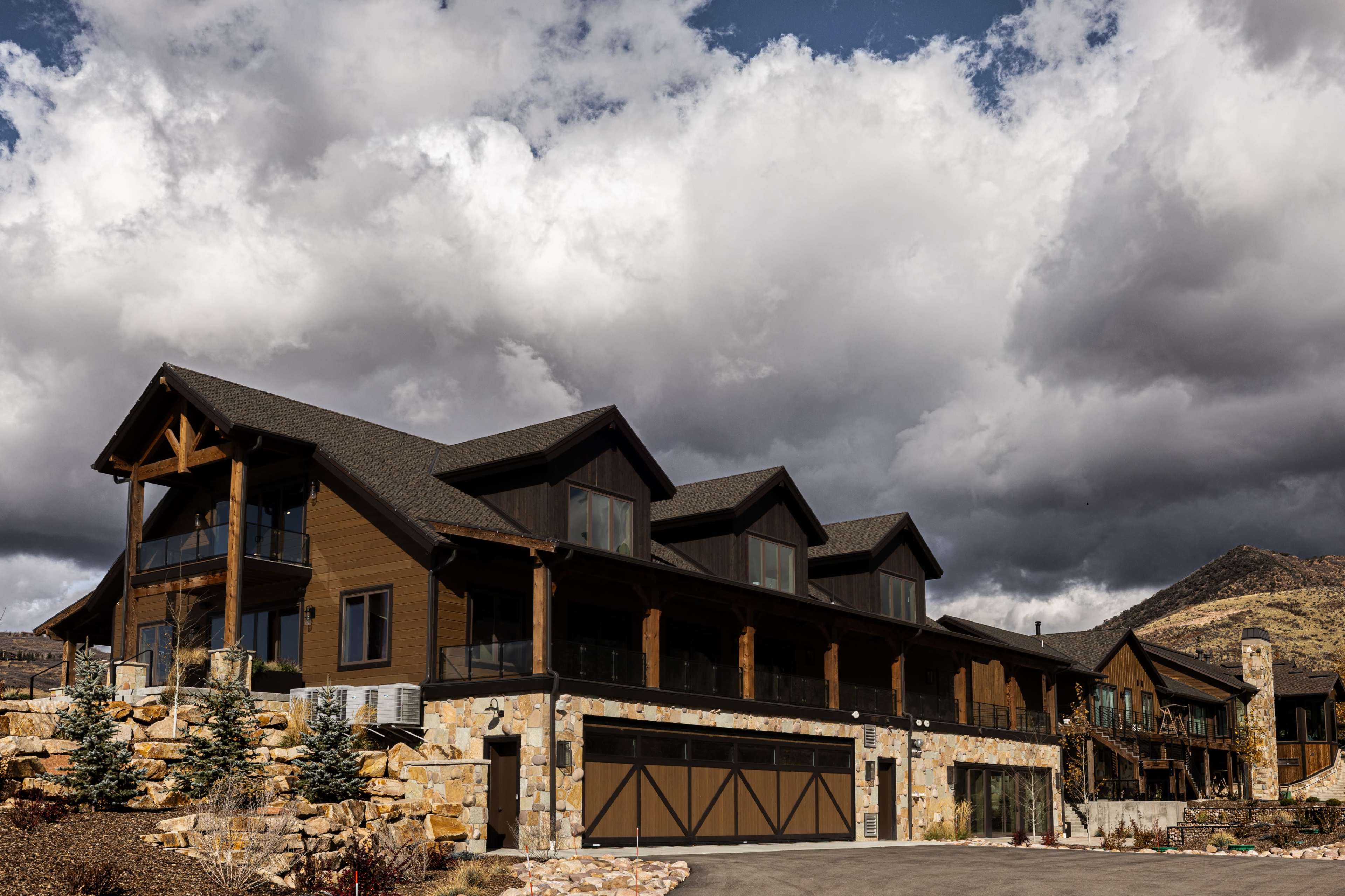 A large, modern mountain home with multiple gabled roofs sits against a backdrop of dramatic clouds and steep hills.