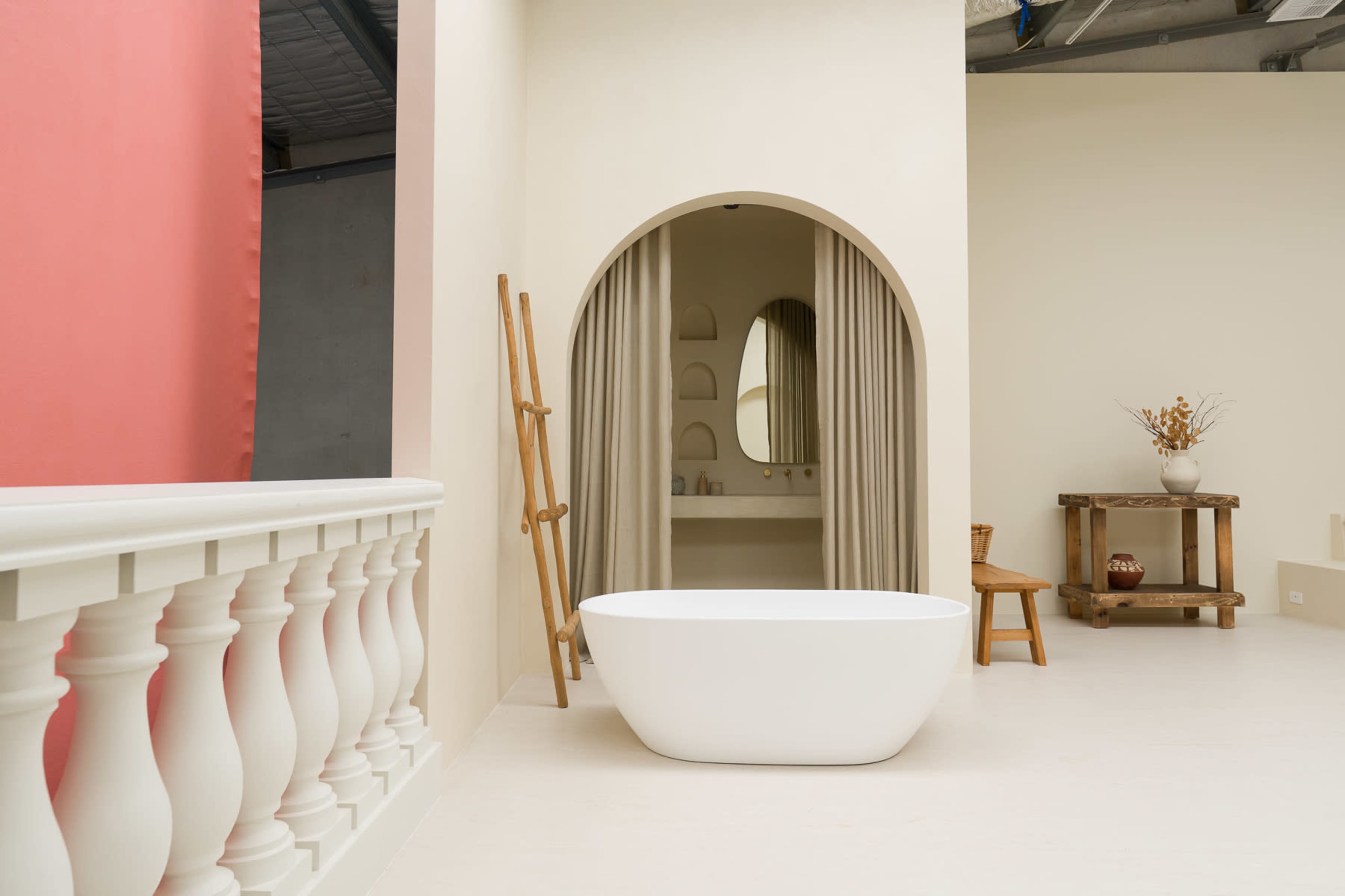 The image shows a modern bathroom with a freestanding white bathtub, a curtained alcove, a wooden ladder, and minimalist decor.