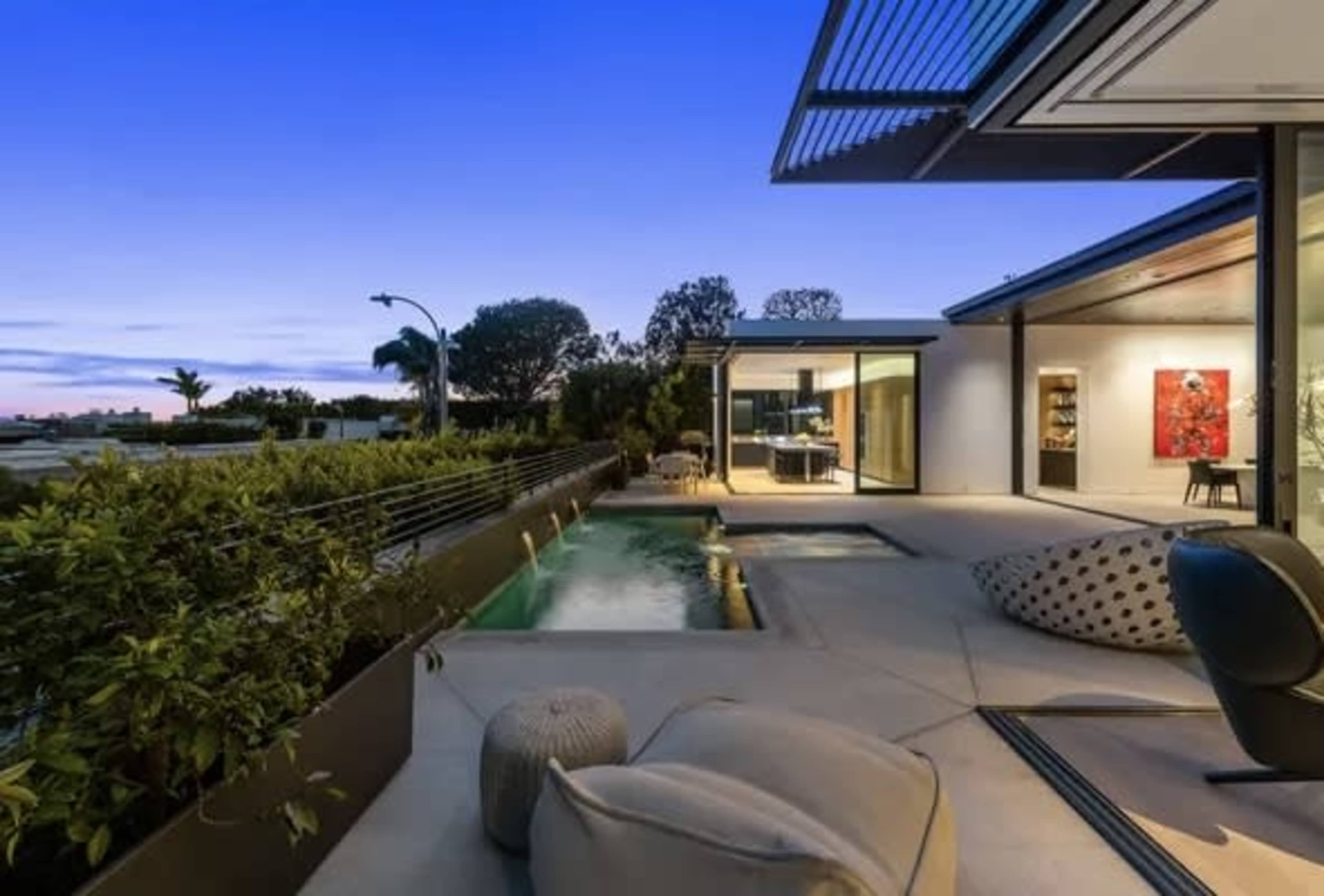 The image shows a modern home with an outdoor living area, featuring a pool, landscaped surroundings, and large windows reflecting a twilight sky.