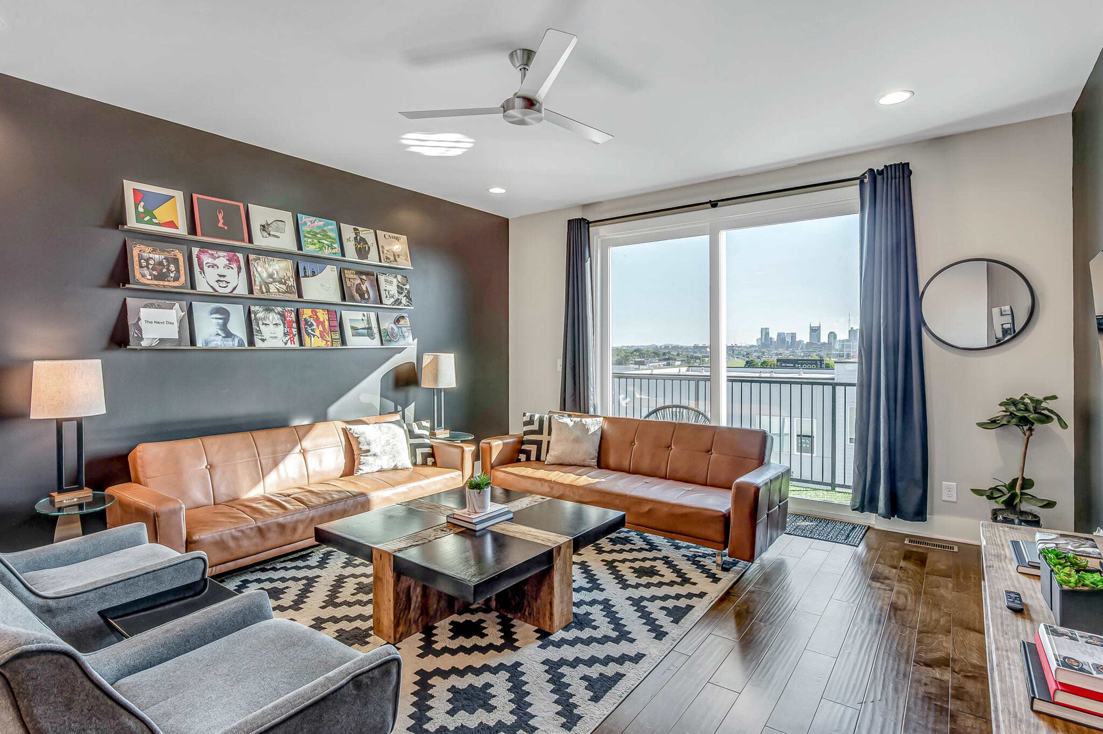 A modern living room features two brown leather sofas, a black coffee table, and a large window with city views, all accented by artwork on the wall and decorative plants.